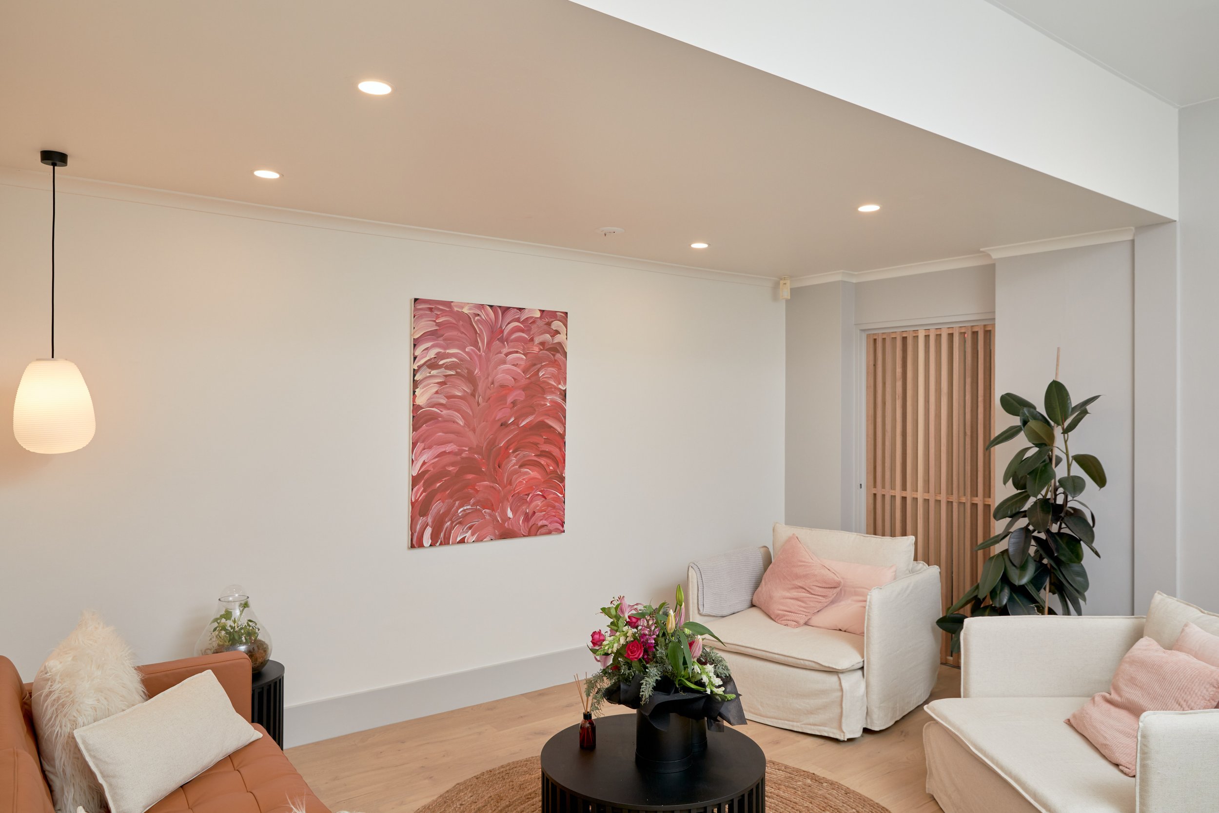 Living room with white walls, beige sofas with pink and white pillows, wooden flooring, a black round coffee table with a flower arrangement, a large potted plant, a wall painting with pink abstract design, hanging pendant light, and a wooden sliding door.