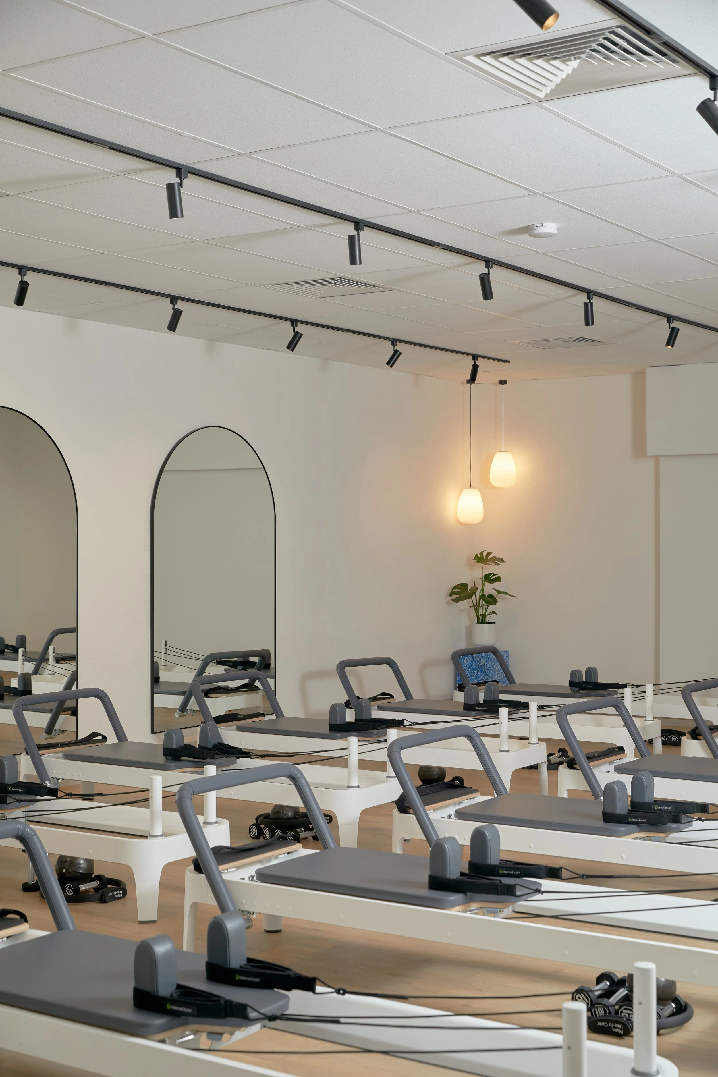Pilates reformer machines in a well-lit studio with mirrors, ceiling track lights, and minimal decor including plants and pendant lights.