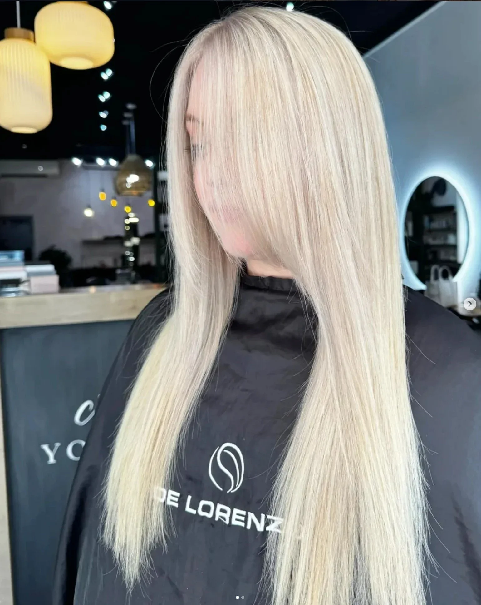 Blonde woman with straight hair sitting in a salon chair, wearing a black cape with the logo 'Corte De Lorenzo' in a modern hair salon.