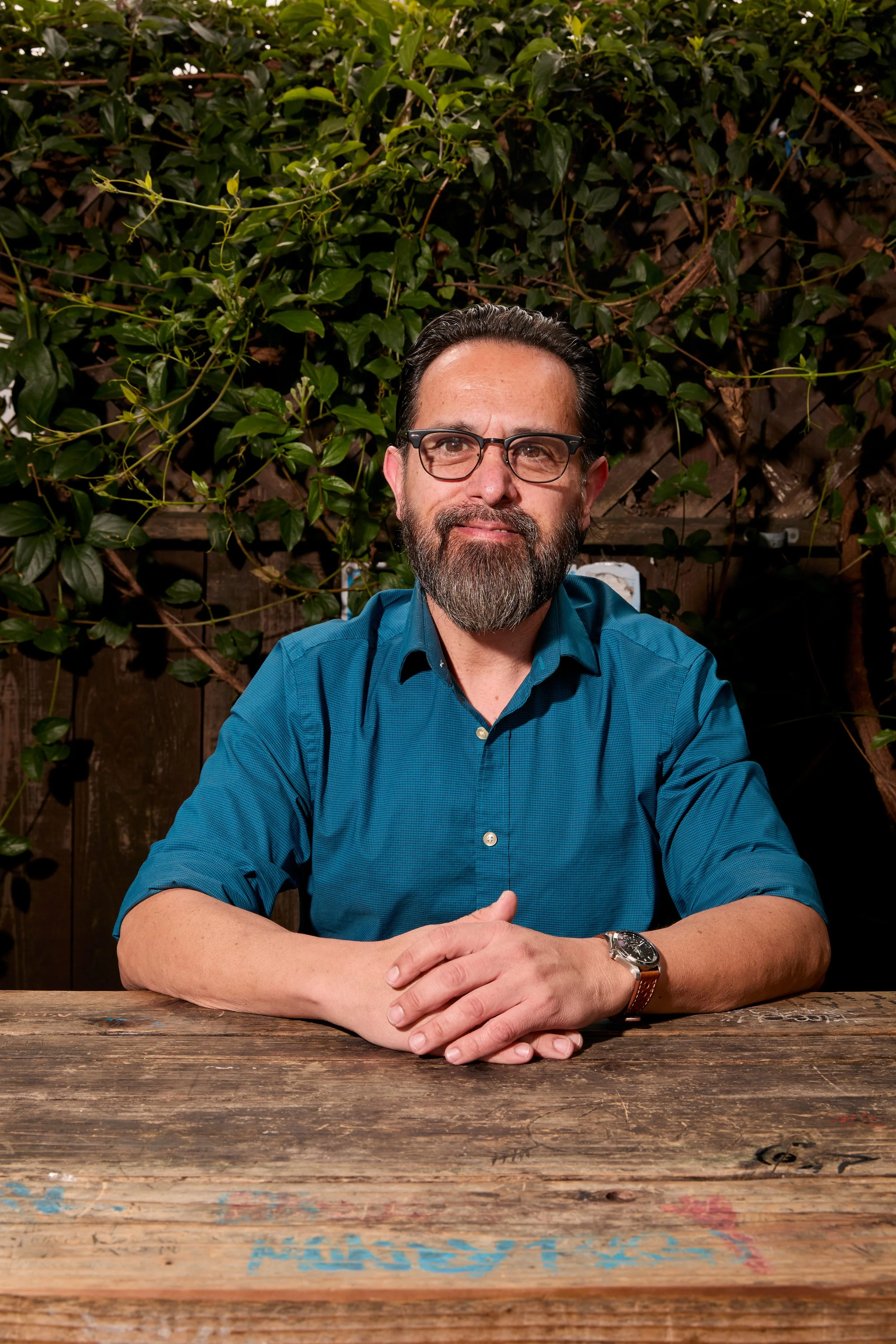 A man with sunglasses and a beard, wearing a light blue shirt and a black apron, sits at a wooden table outdoors with a background of leafy green plants and pink flowers.