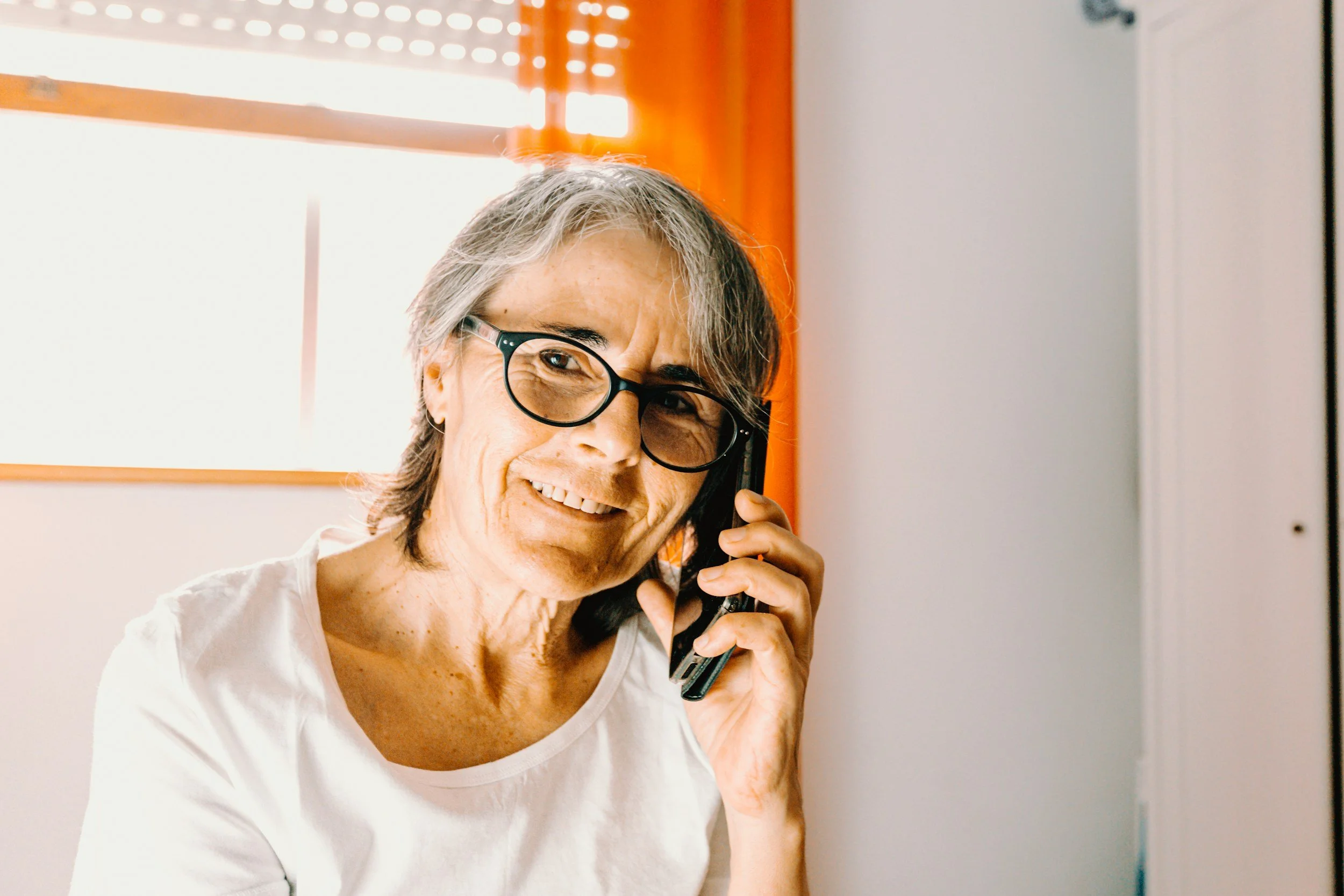 An elderly woman with glasses and grey hair talking on a mobile phone, smiling in a brightly lit room with orange curtains and a white wall.