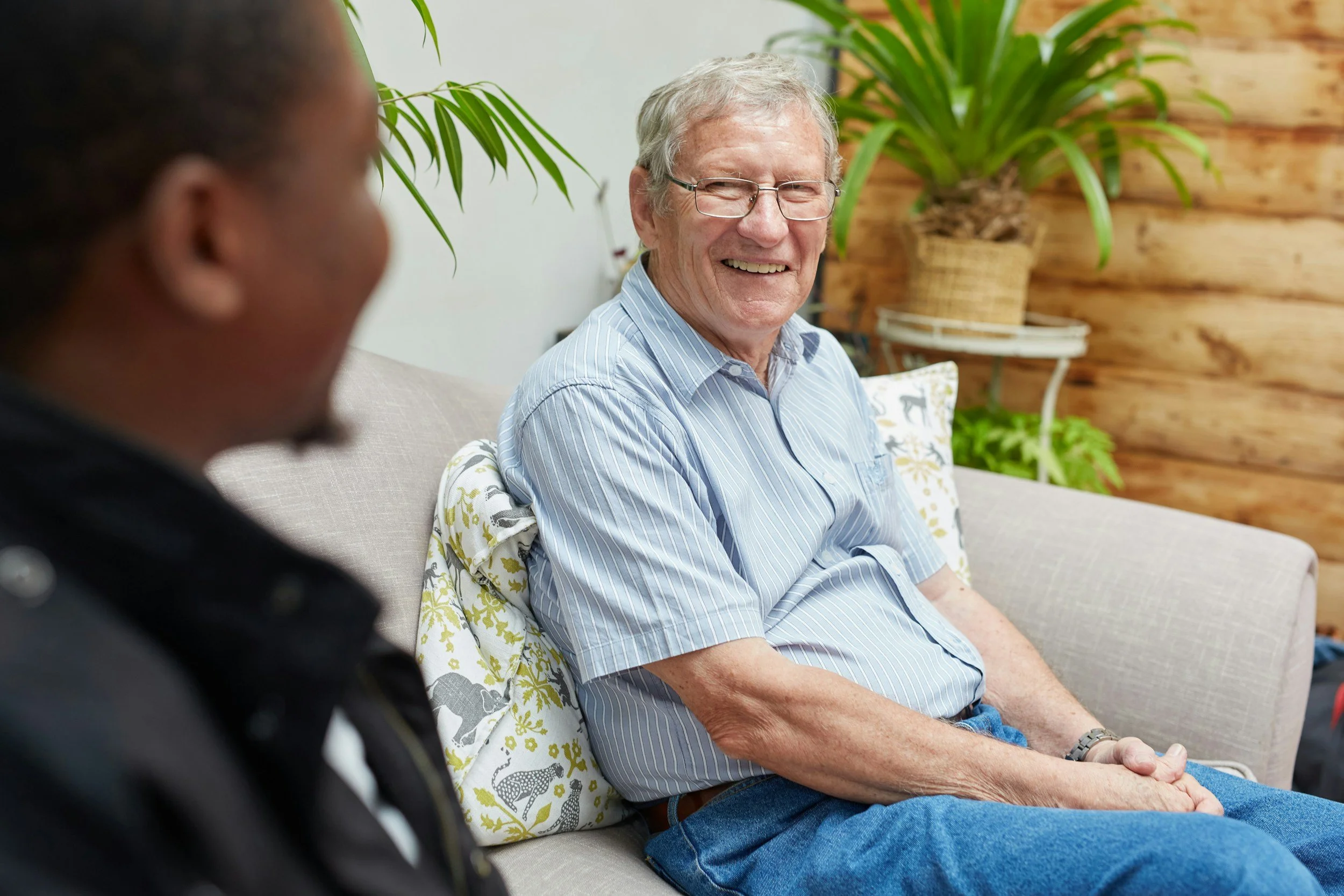 An elderly man with glasses, smiling, sitting on a beige couch with a pillow, engaging in conversation with a person out of focus in the foreground, in a cozy room with wooden walls and green indoor plants.