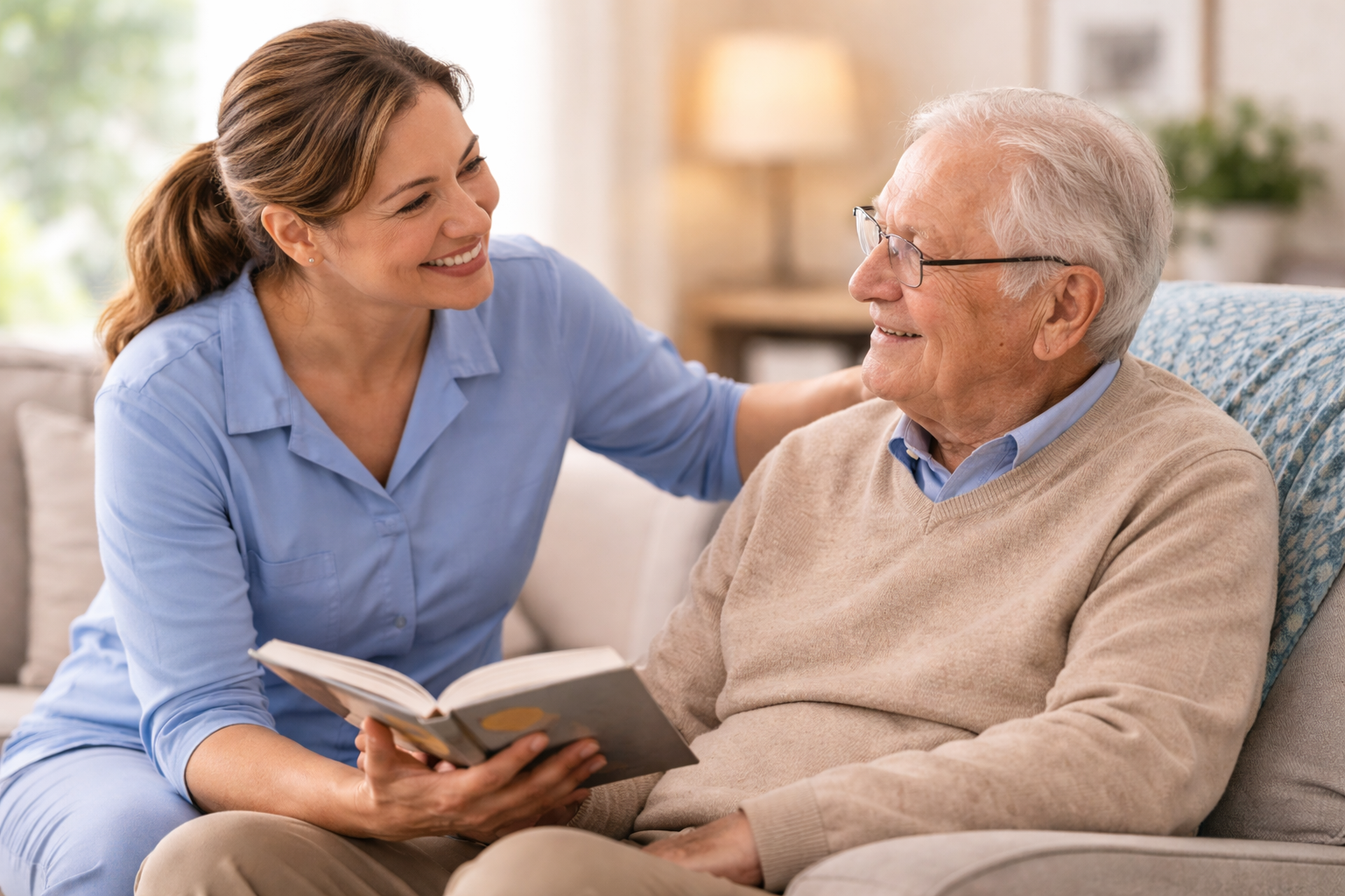 A young female caregiver smiling and talking to an elderly man with glasses, holding a book, sitting on a couch in a living room.