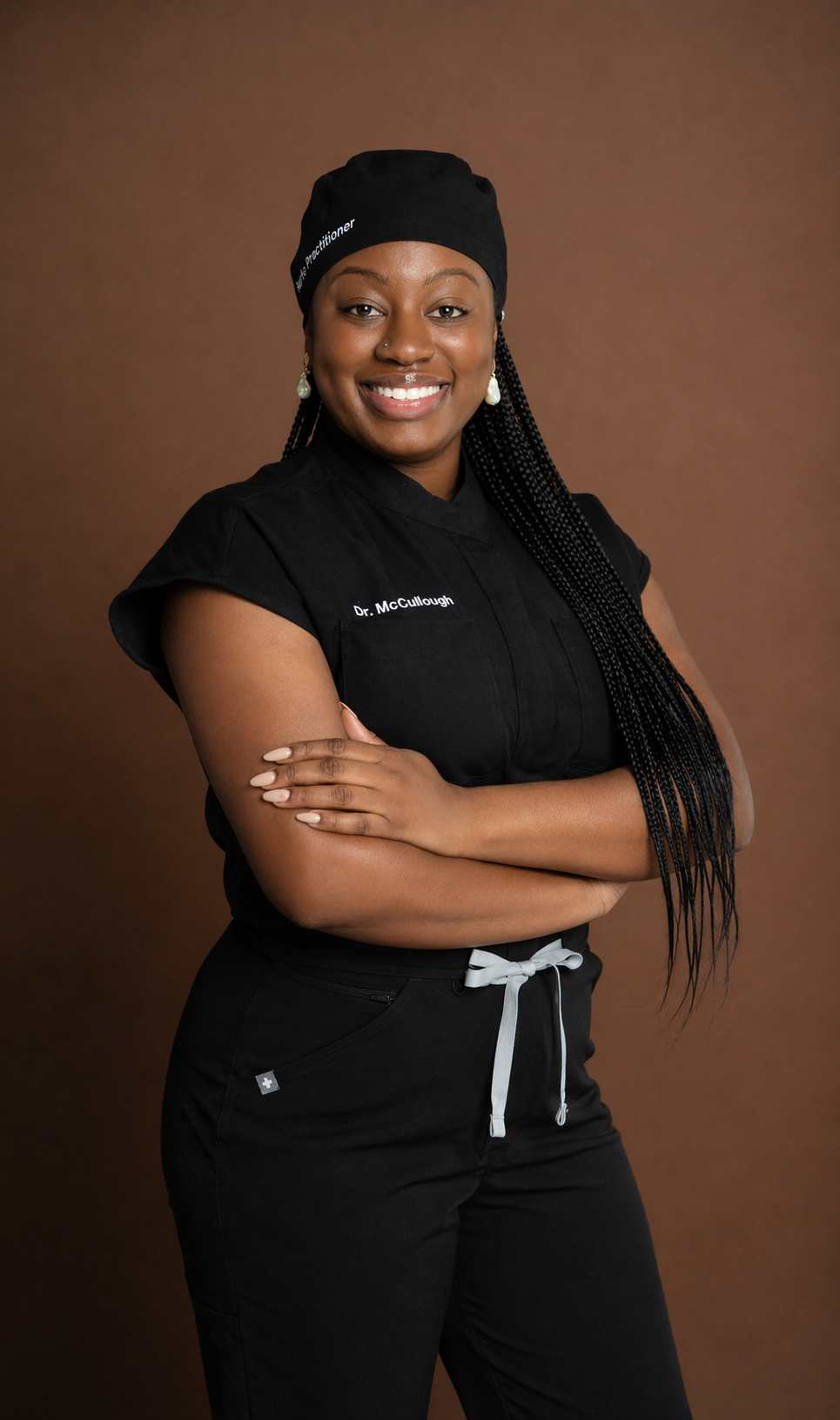 Professional woman in medical scrubs with crossed arms, wearing a black head covering and earrings, smiling in front of a brown background.