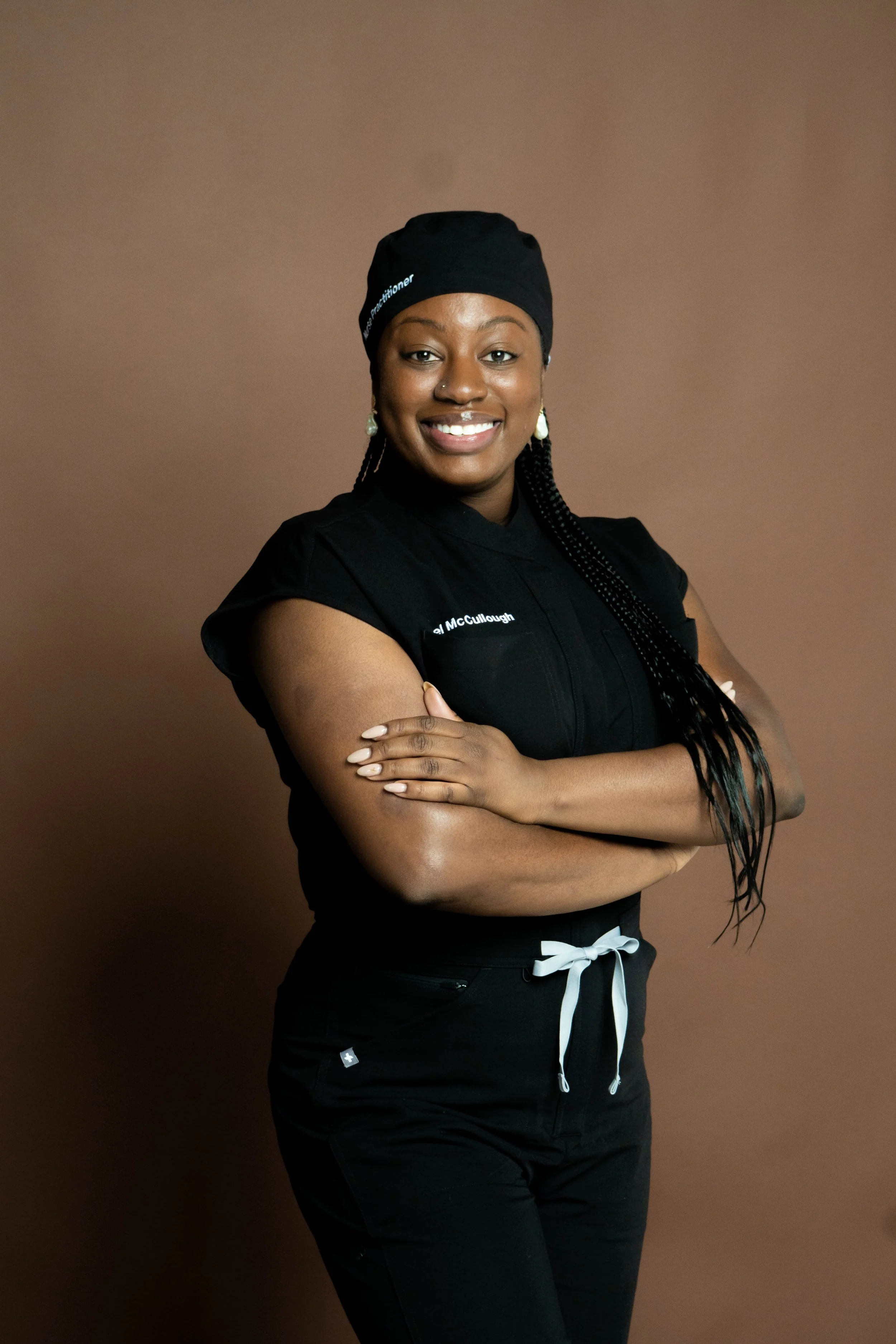 A woman with braided hair and earrings, dressed in black scrubs with a black cap, standing with arms crossed and smiling against a brown background.