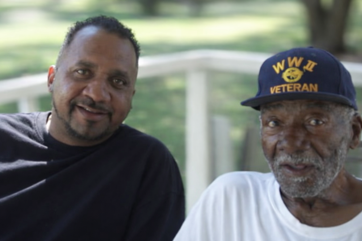 A middle-aged man and an elderly man sitting outdoors on a bench. The middle-aged man is smiling and has short curly hair, wearing a black T-shirt. The elderly man is wearing a navy cap with yellow text and a yellow emblem, and a white T-shirt. The background shows greenery and a white railing.
