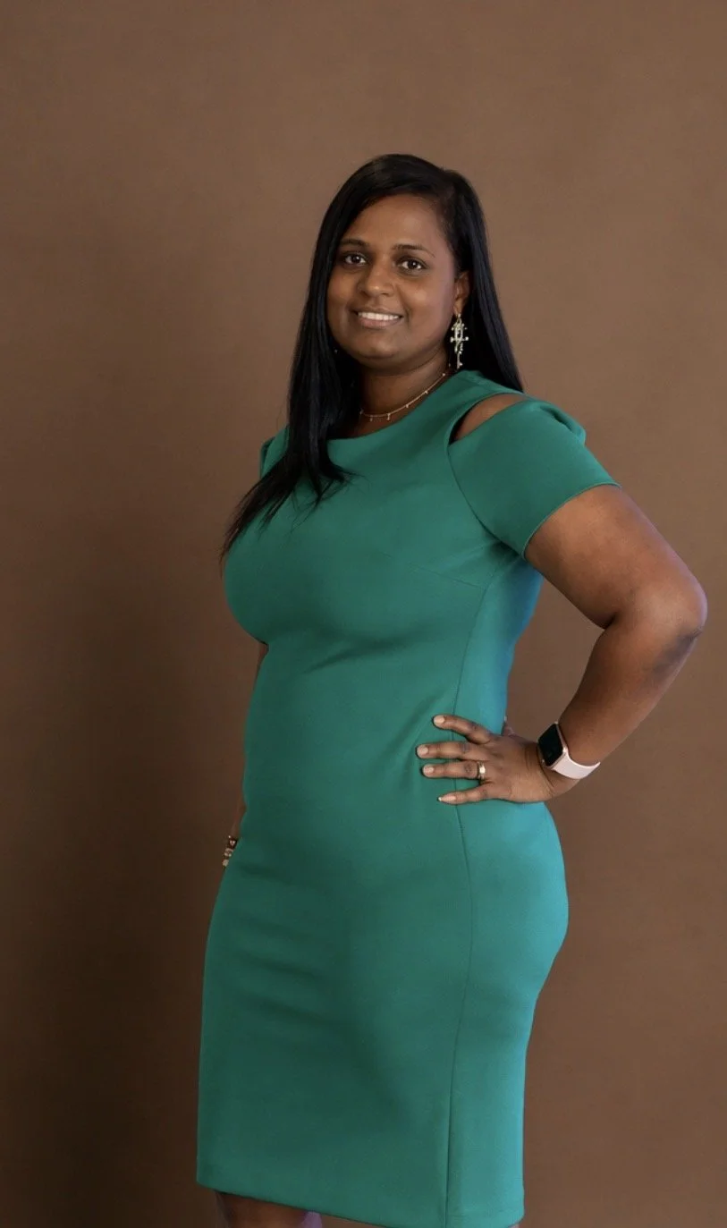 A woman with dark hair and medium skin tone, wearing a teal dress with cutout shoulder details, standing against a brown background.