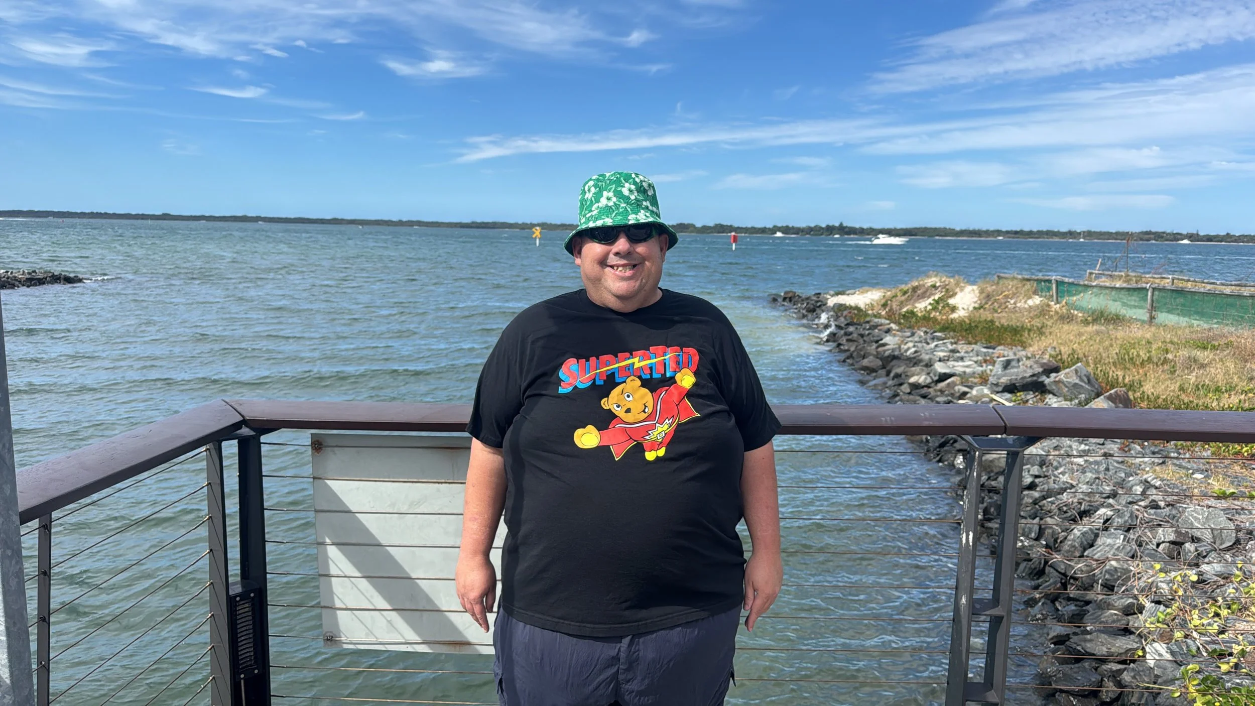A man wearing a green floral bucket hat, sunglasses, a black T-shirt with a cartoon character and the word 'SuperTed,' and gray shorts, standing on a railing near the water on a sunny day with blue sky and clouds.