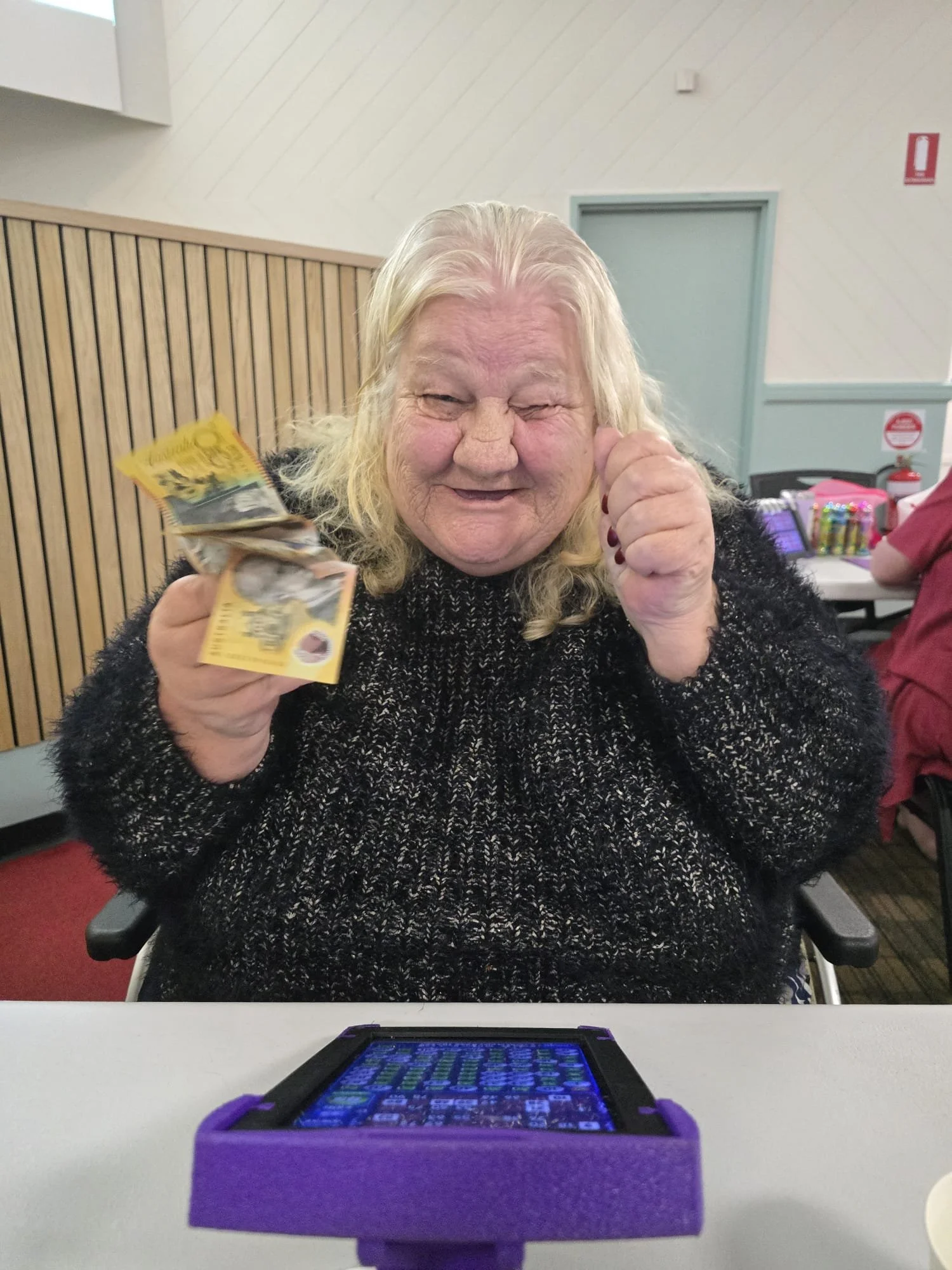 An elderly woman with long blonde hair, wearing a black sweater, smiling cheerfully while holding dollar bills in her hand. She is sitting at a table in a room with white walls and wooden paneling, with a tablet device in a purple case in front of he