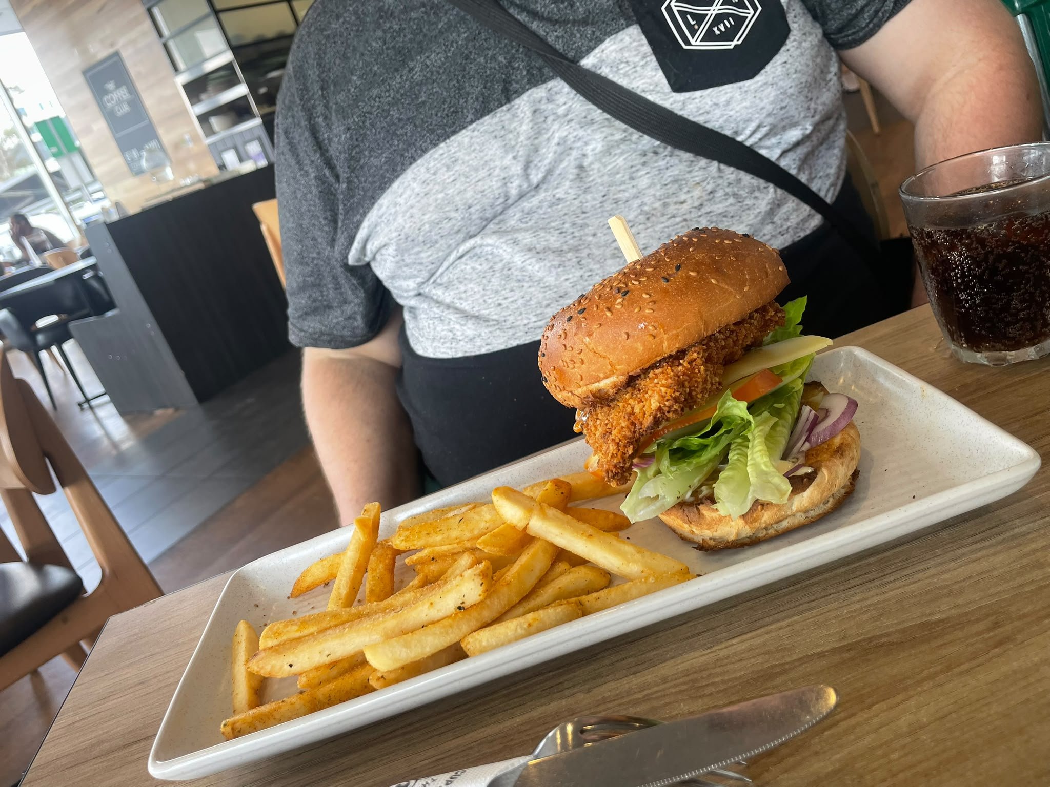 A person sitting at a table in a restaurant with a tray of food in front of them. The tray contains a fried chicken sandwich with lettuce, tomato, and pickles, topped with a sesame seed bun, alongside a portion of seasoned French fries. A glass of da