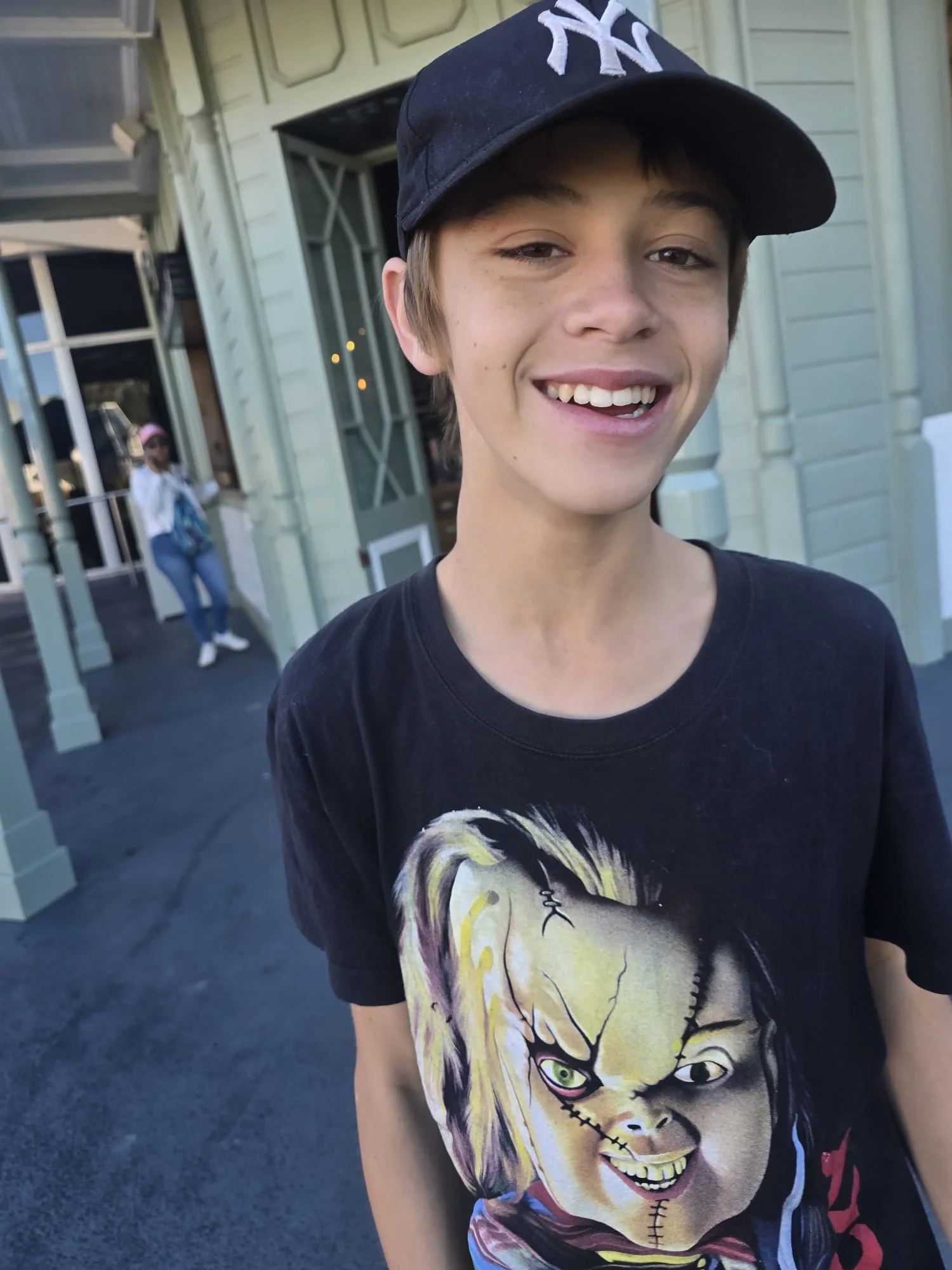 Close-up of a young person smiling, wearing a black New York Yankees cap and a graphic t-shirt featuring a cartoon character with blonde hair and a stitched mouth.