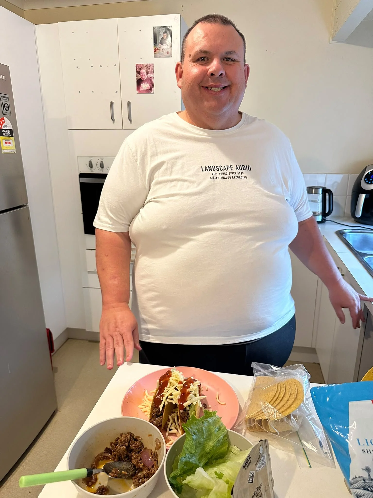 A man standing in a kitchen smiling at the camera, with a table in front of him filled with dishes including a plate of spaghetti with chili, a bowl of seasoned ground beef and onions, a salad with lettuce, and snacks like cookies and potato chips.