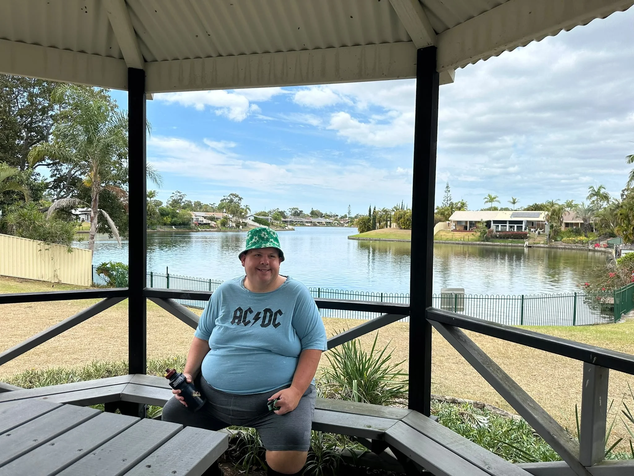 A smiling person wearing a blue AC/DC t-shirt and a green hat with white patterns, holding a drink, sitting on a gazebo with a lake and houses in the background.