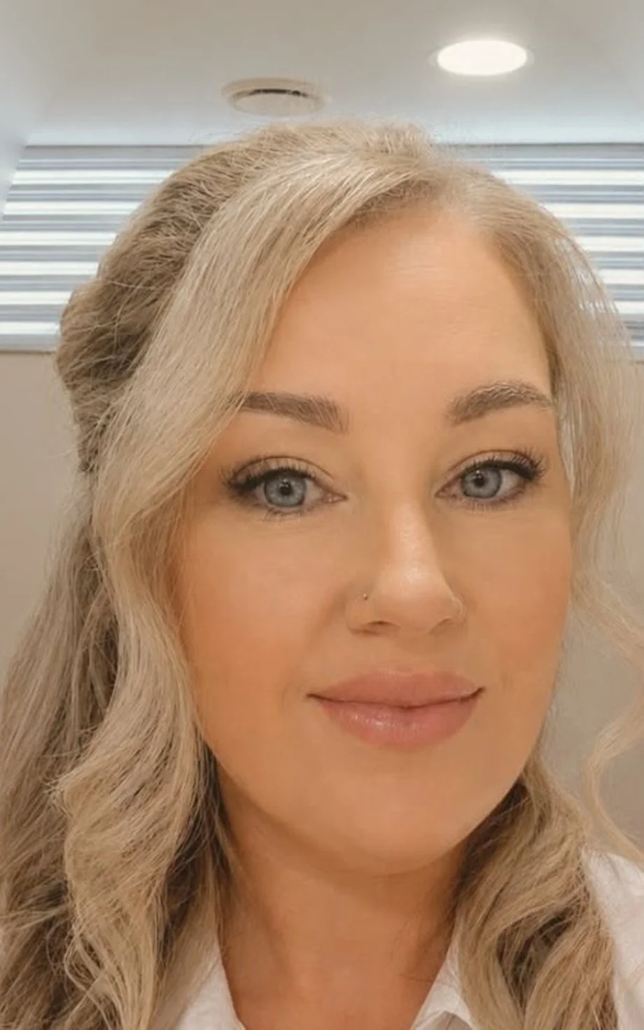 A woman with long blonde hair, blue eyes, and light makeup, wearing a white top, smiling slightly in front of a window with horizontal blinds.
