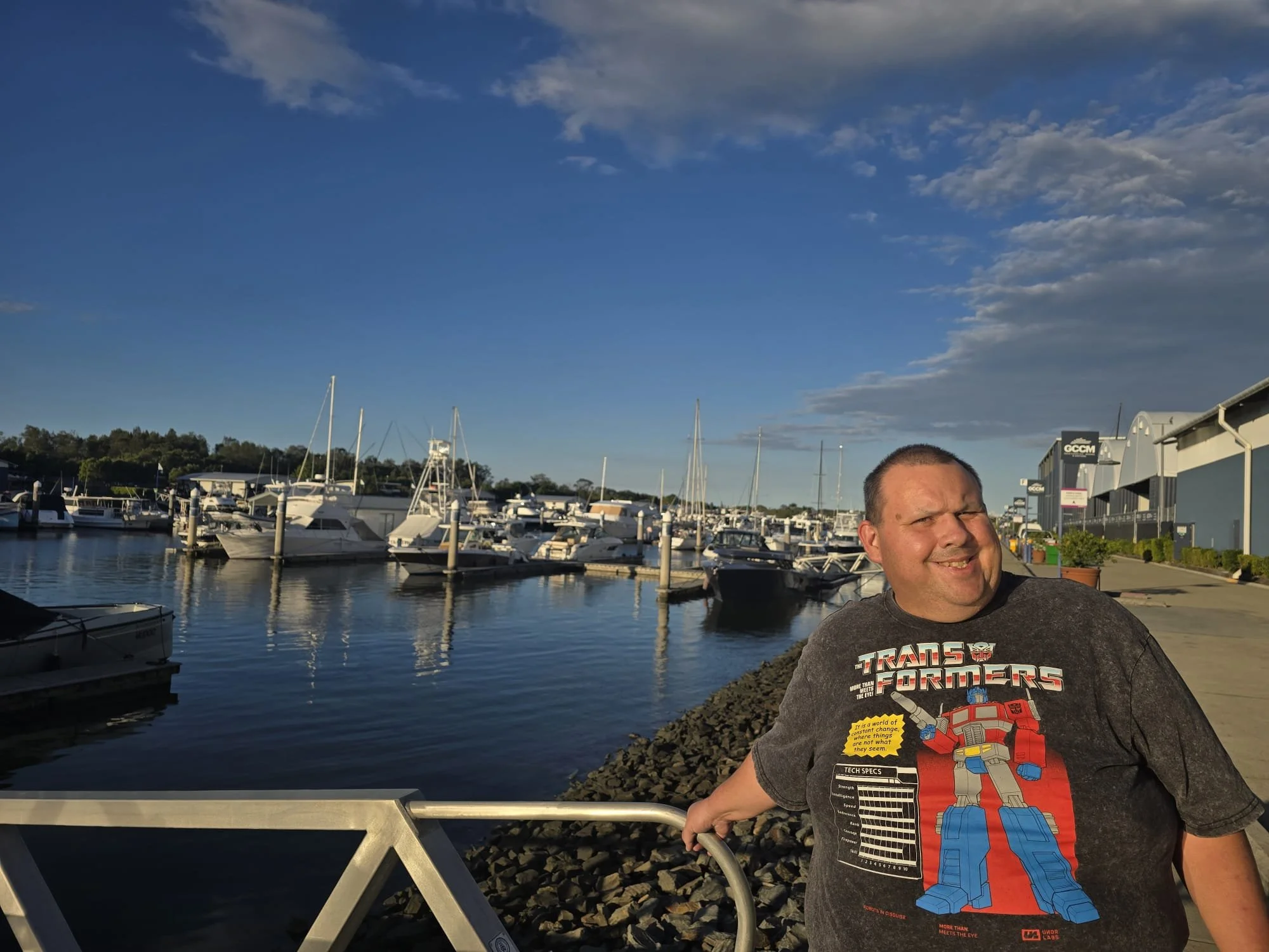 A man smiling at the camera, standing outdoors near a marina with boats docked in the background, under a partly cloudy sky.