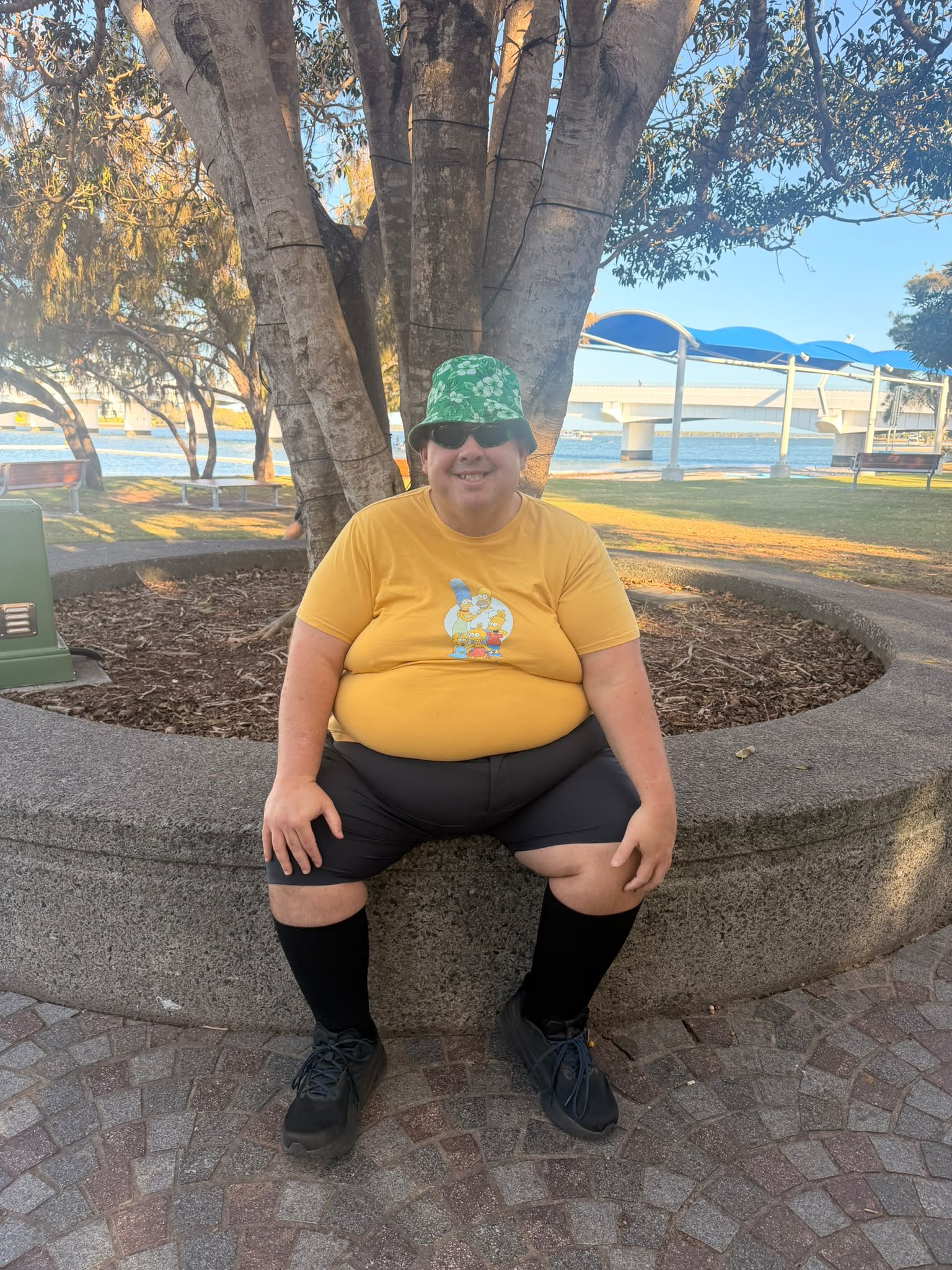 A person sitting on a concrete bench in a park near water, wearing a yellow t-shirt with cartoon characters, black shorts, black shoes, sunglasses, and a green bucket hat with a white floral pattern, with a large tree and a bridge in the background.