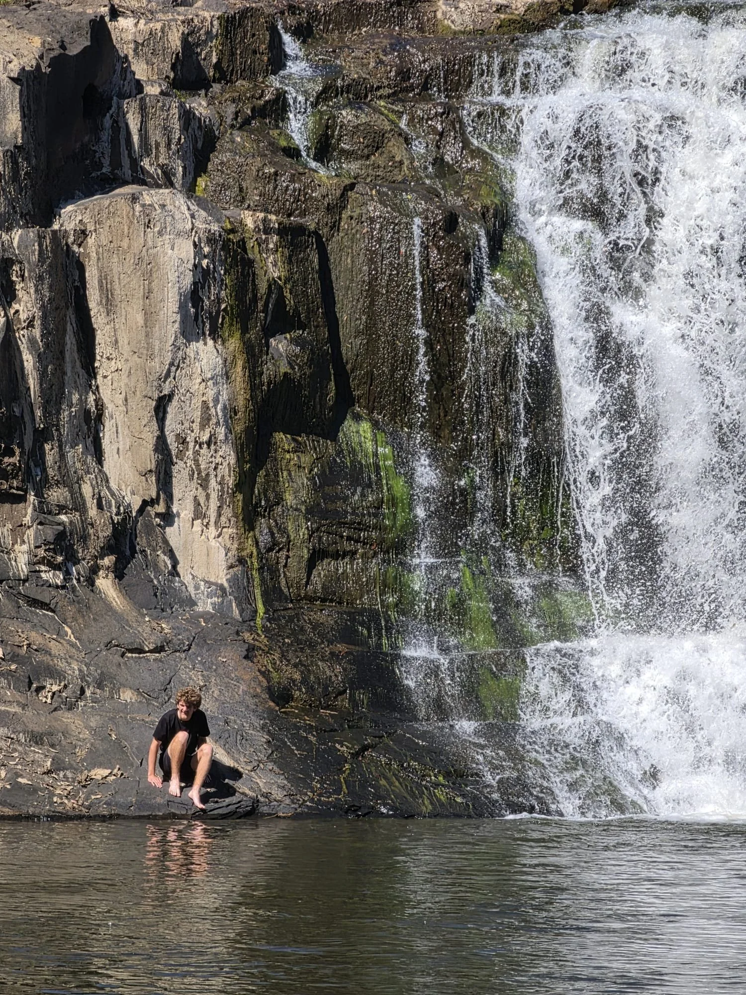 A person sitting on rocks near a waterfall with water cascading over dark rocks and green moss, reflecting in a calm pool.