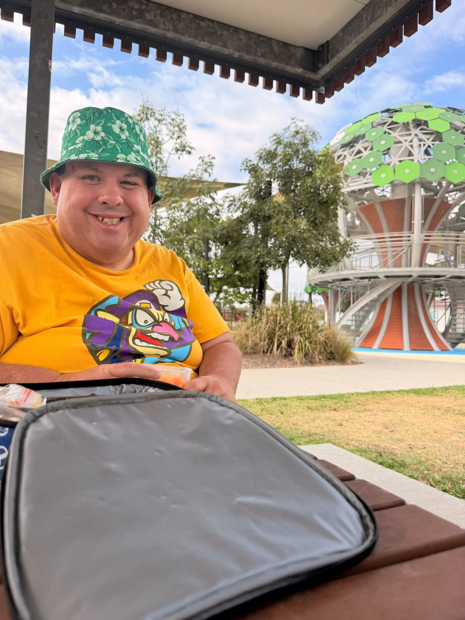A man with a wide smile, wearing a green floral bucket hat and a bright yellow T-shirt with a colorful graphic, sitting at a picnic table outdoors at an amusement park or fairground with a large Ferris wheel and trees in the background.