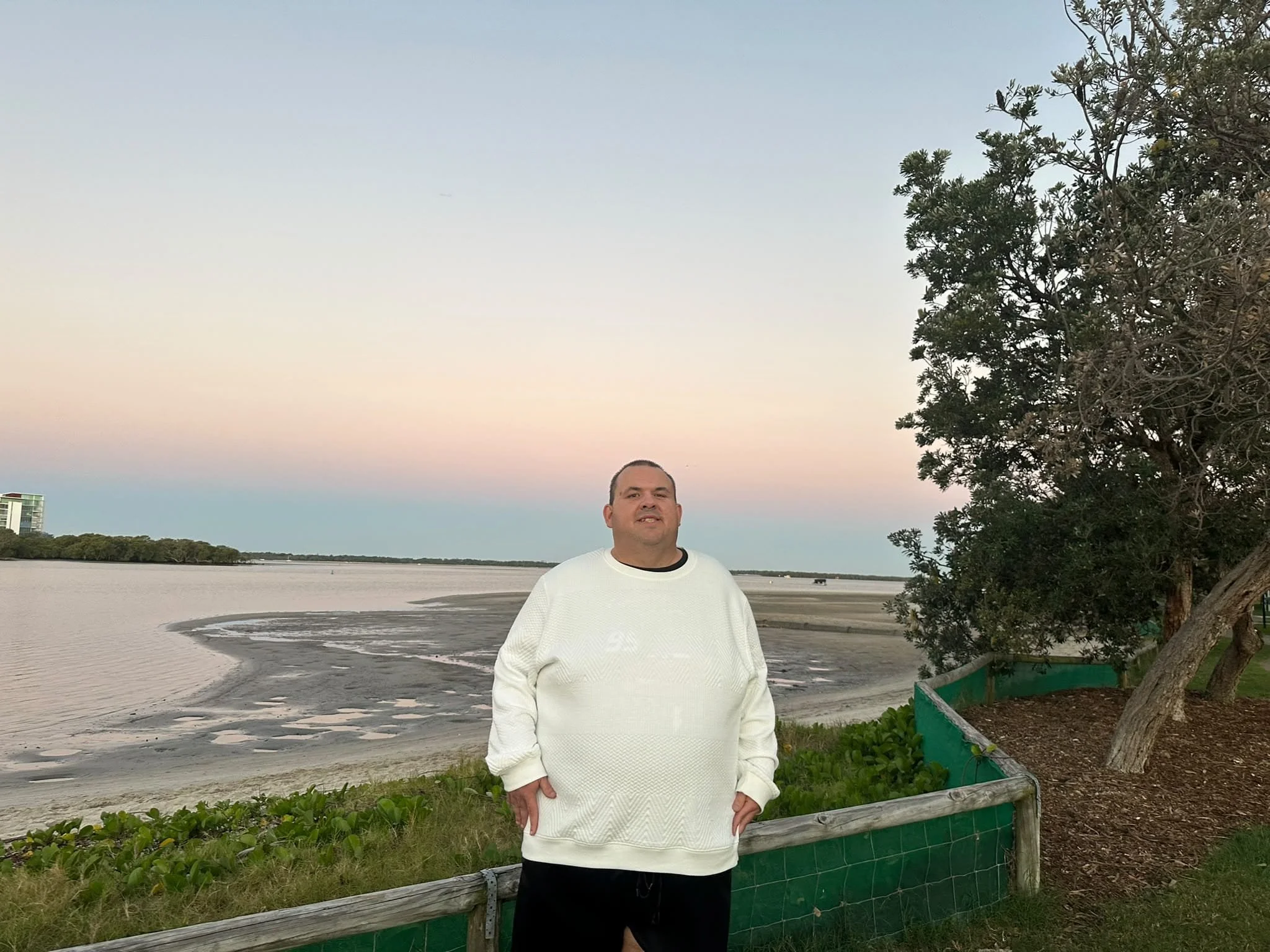 A man standing outdoors by a river or bay, wearing a white sweater and black shorts, with trees and water in the background during what appears to be sunset or sunrise.