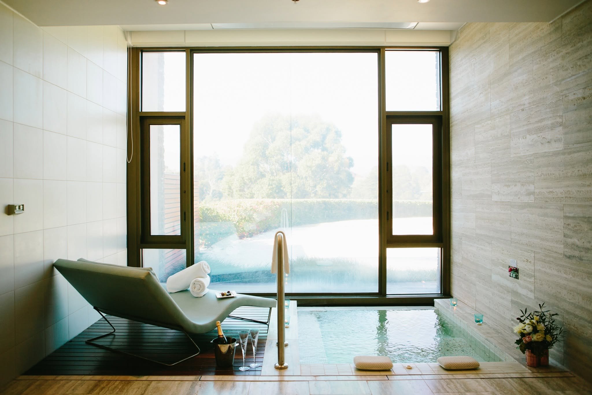 Indoor spa room with a view of trees through large floor-to-ceiling windows, a lounge chair with towels, champagne glasses, and a flower arrangement.