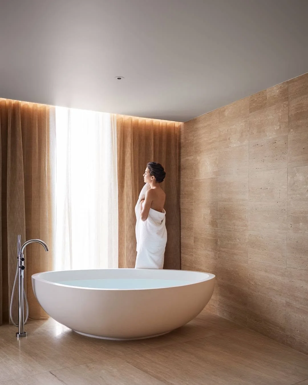 A woman wrapped in a white towel standing in a modern bathroom, looking out of a large window with beige curtains, next to a freestanding bathtub.