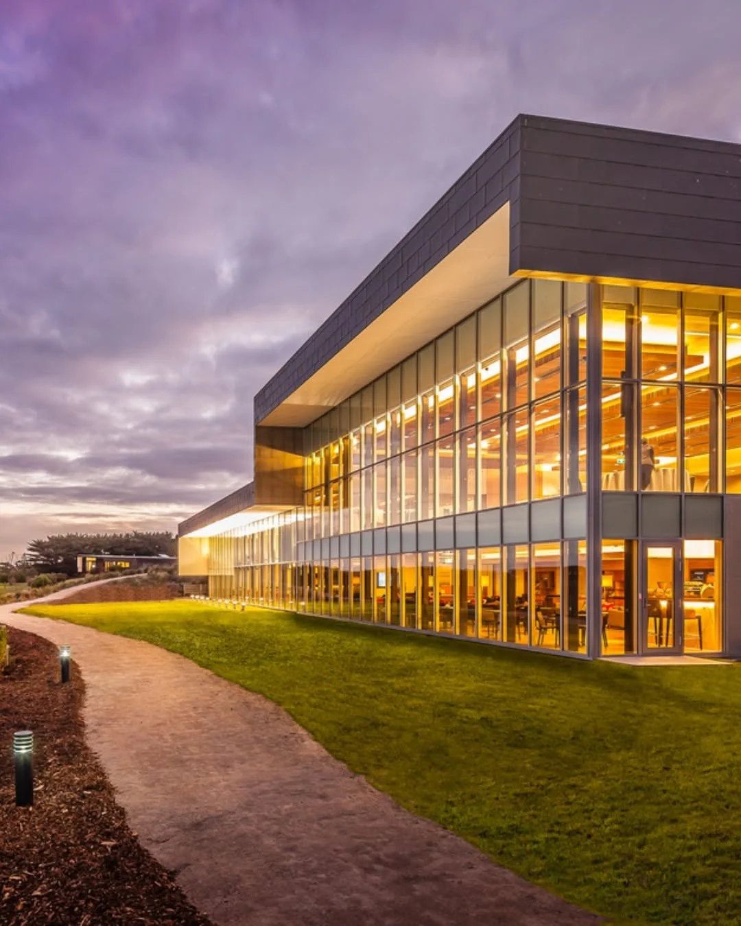 Modern two-story office building with glass exterior illuminated from inside, surrounded by a grass lawn and a paved walkway, under a partly cloudy evening sky.