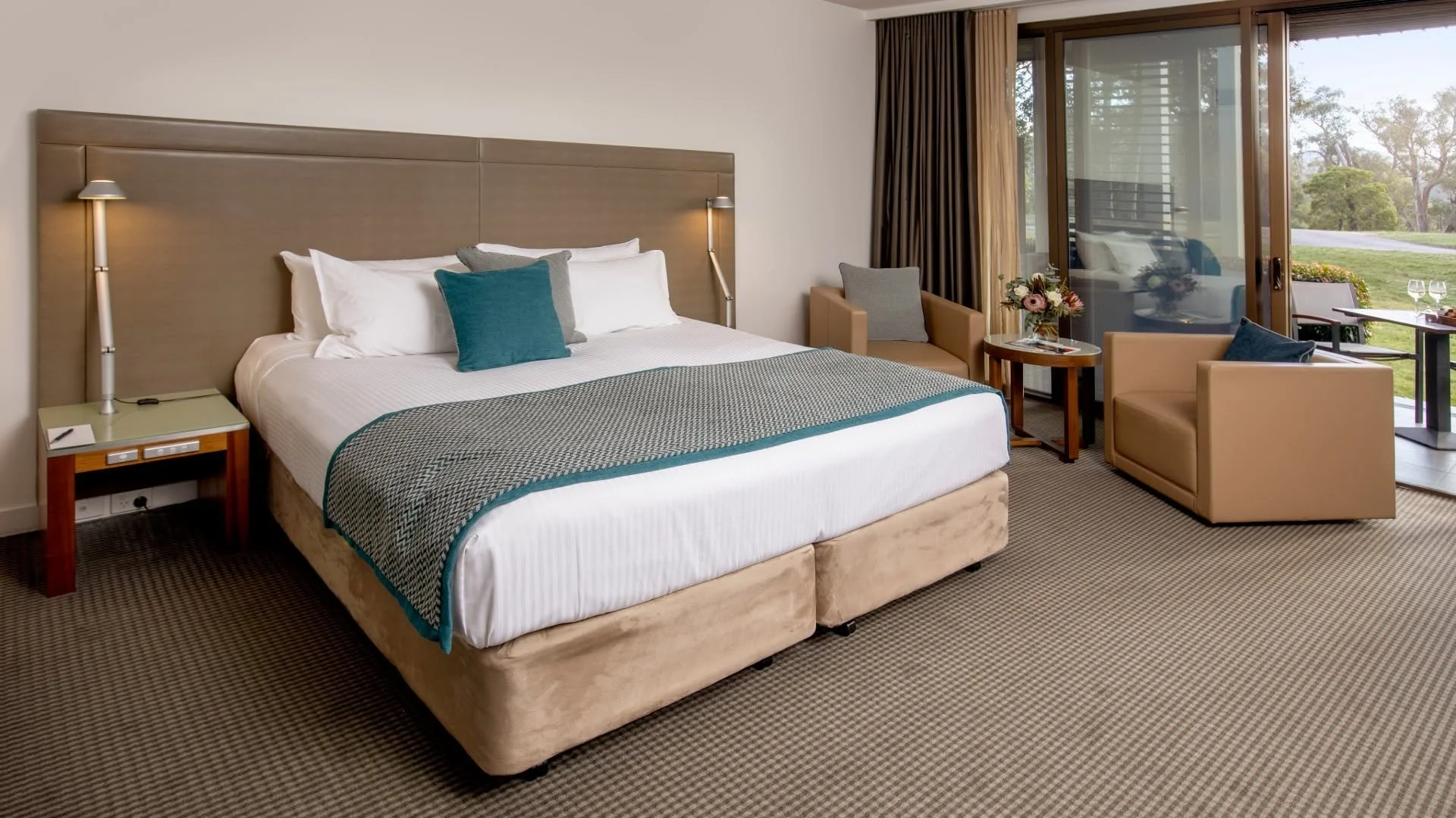 A hotel room with a large bed, bedside tables, and a sitting area with two armchairs and a small round table. Sliding glass door opens to a balcony with a view of trees and a grassy area.