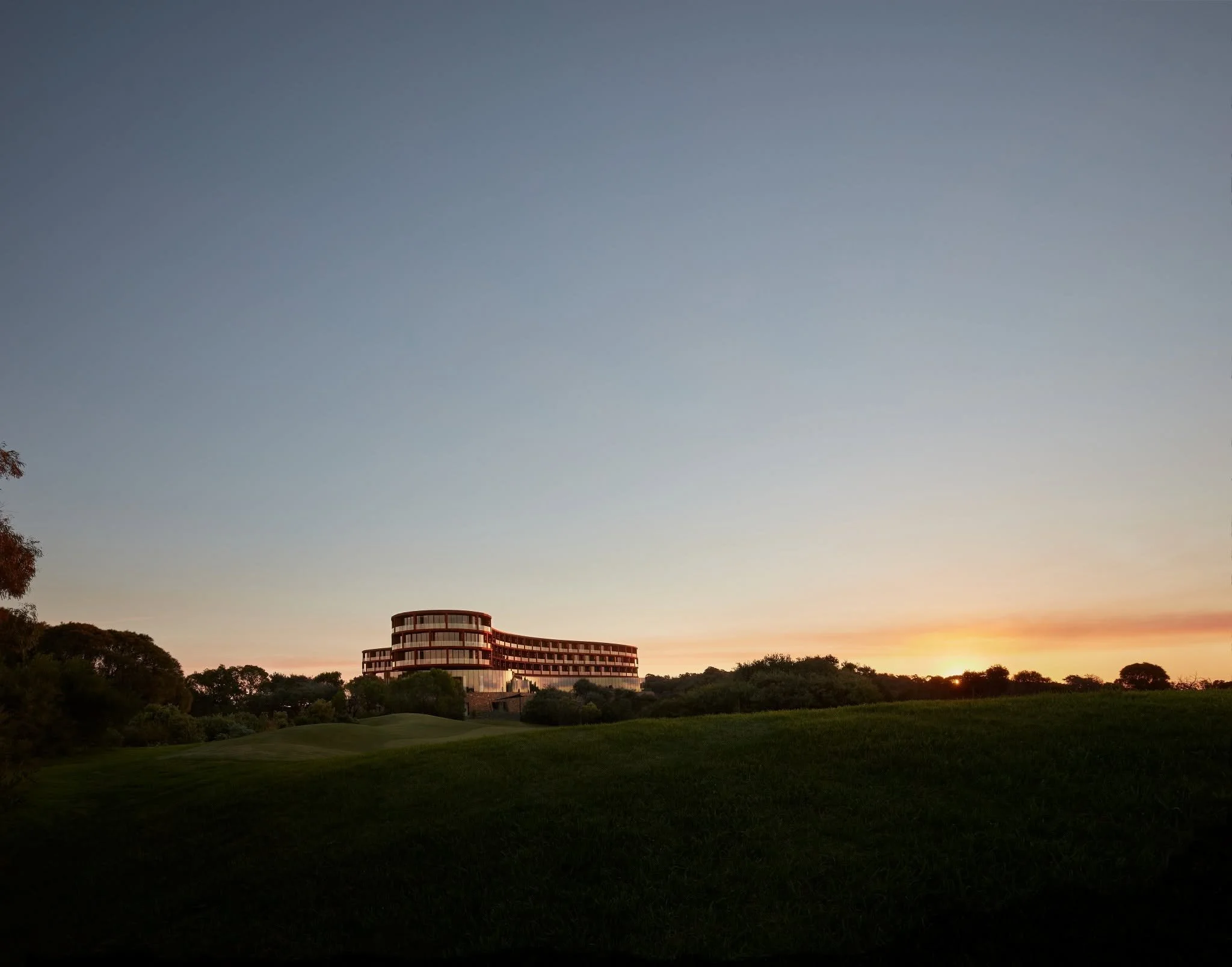 A curved modern building with multiple floors, set amidst a lush green landscape, under a twilight sky