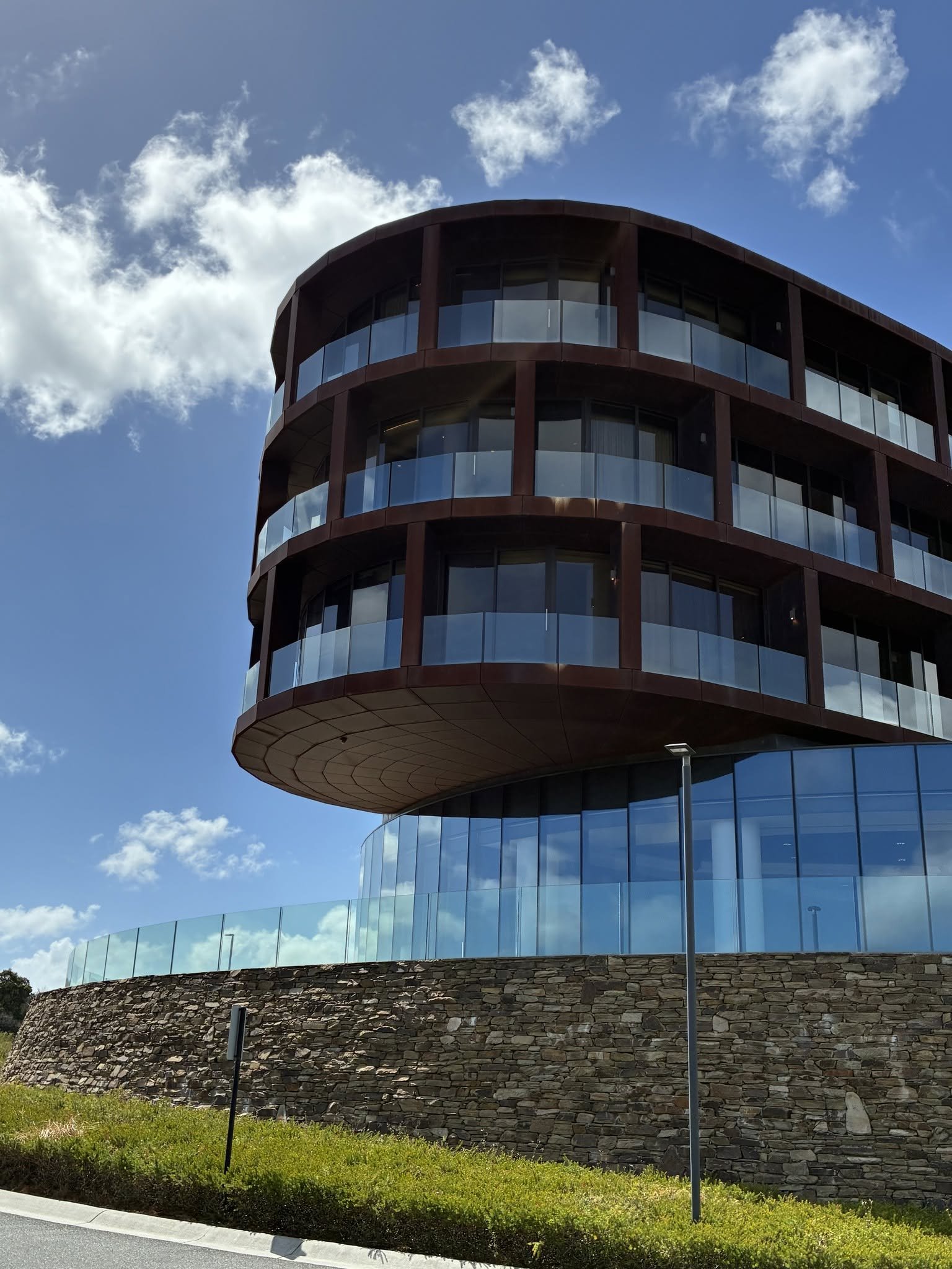 Modern multi-story building with curved glass balconies and a stone base, against a blue sky with clouds.