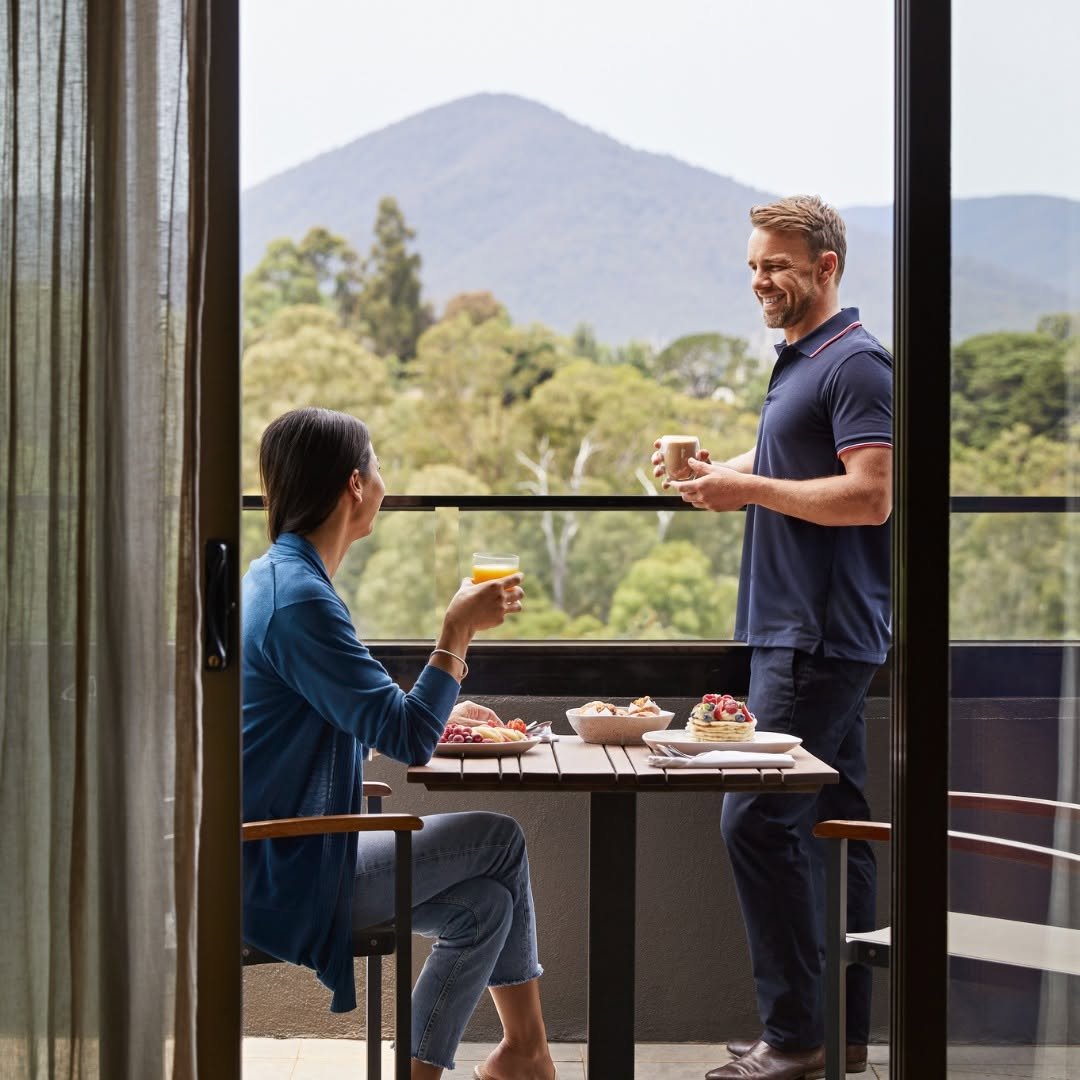 A man and woman on a balcony having breakfast and drinks, with mountains and trees in the background.
