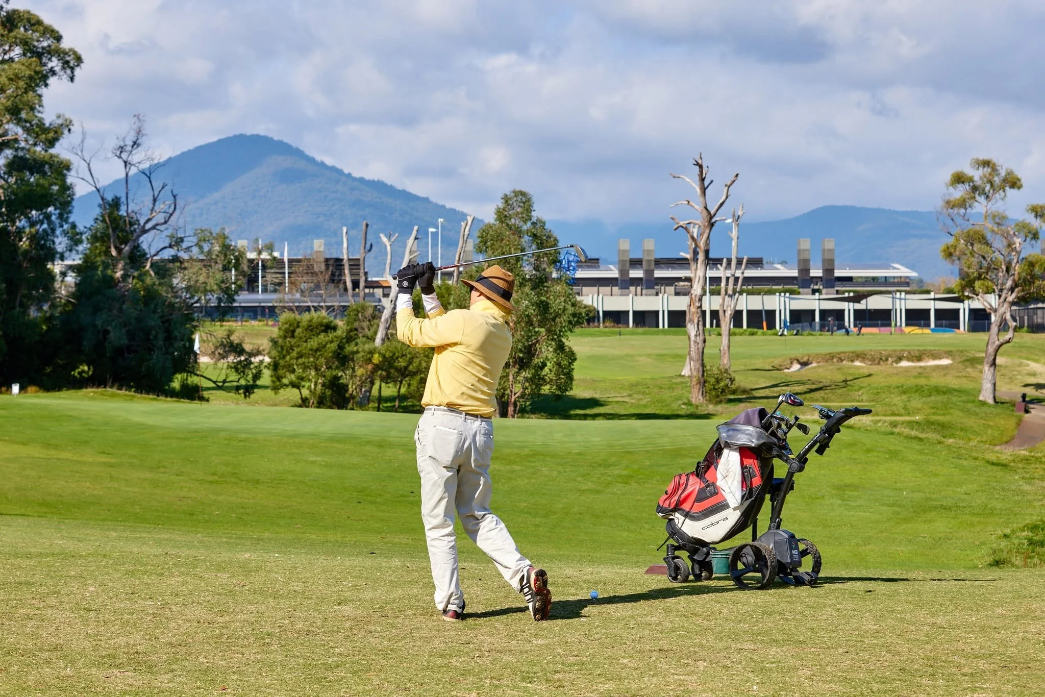 A person in a yellow jacket and beige pants is playing golf on a lush green course, swinging a golf club with a golf bag and cart nearby. The background features trees, modern buildings, and mountains under a partly cloudy sky.