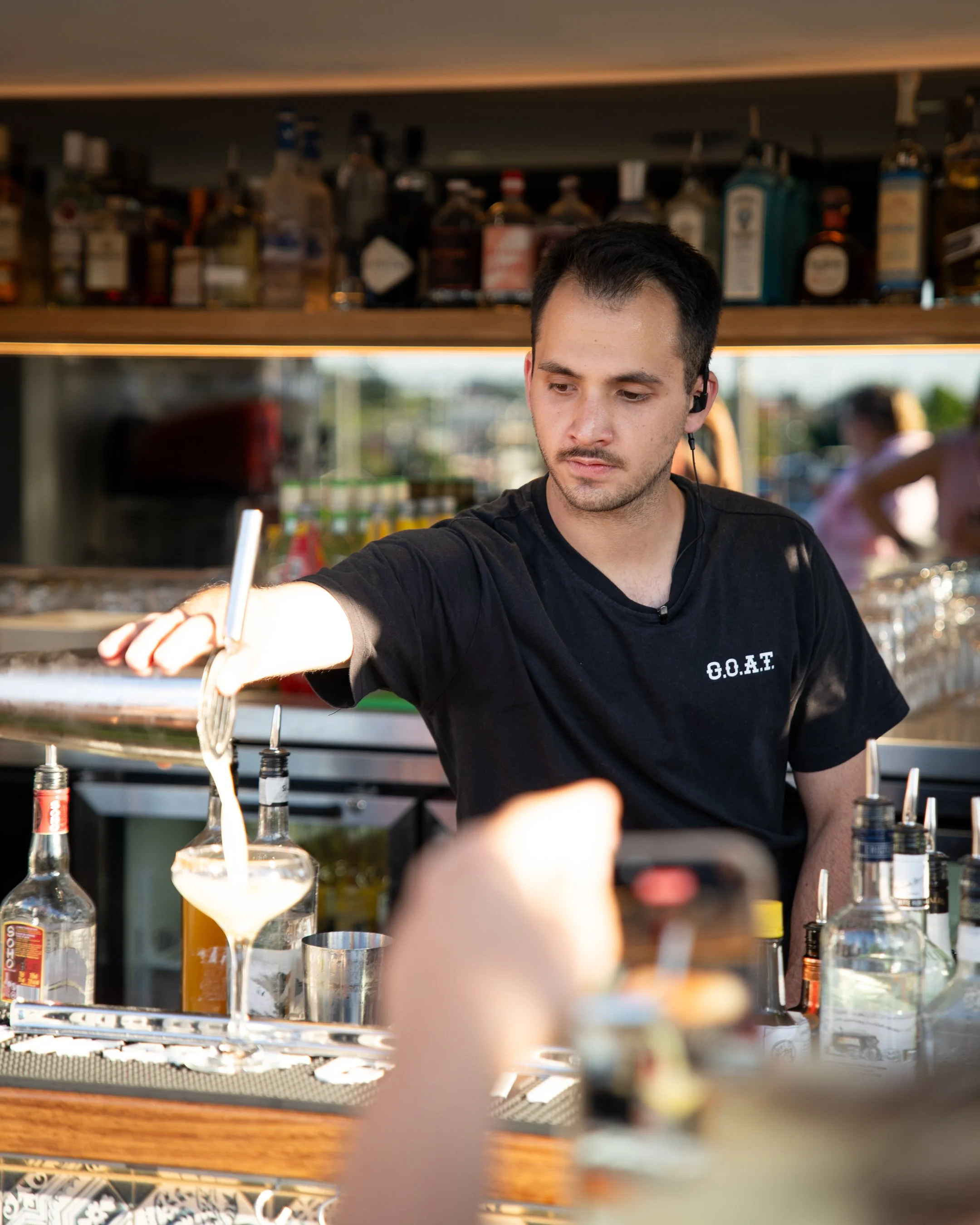 Bartender pouring a drink at a bar with liquor bottles in the background.