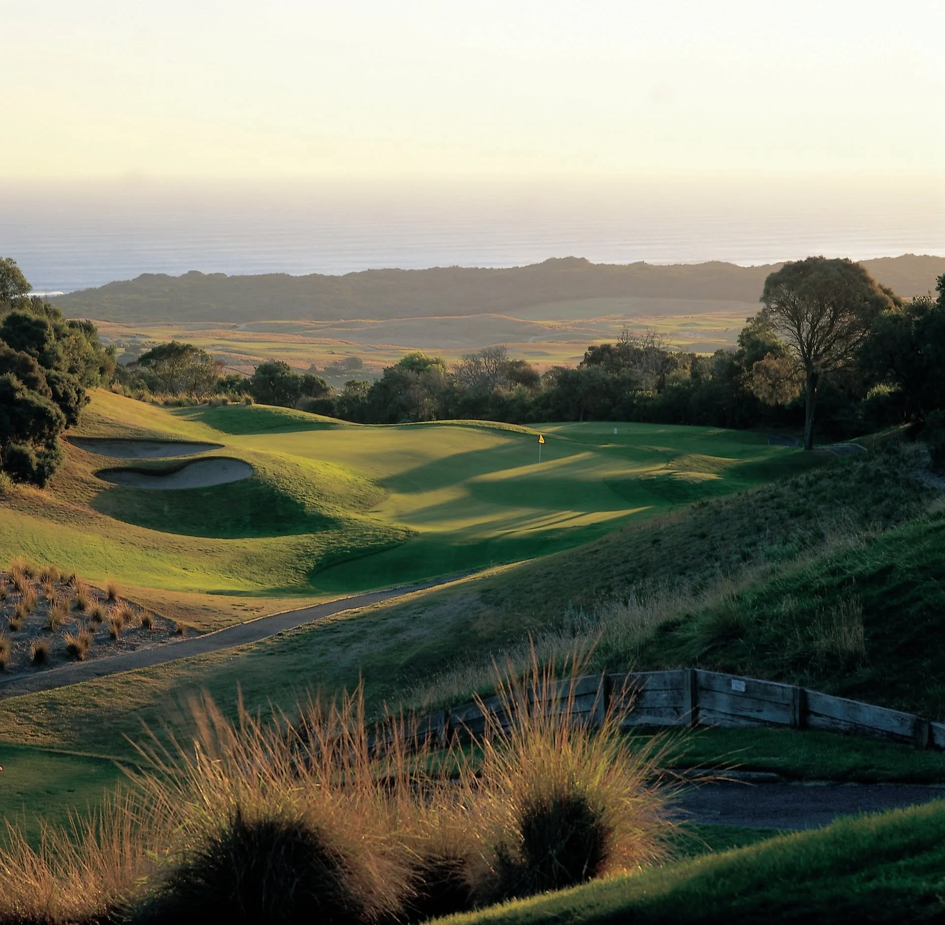 Landscape view of a golf course with green fairways, sand bunkers, and trees, overlooking a hillside and the ocean in the distance at sunset.