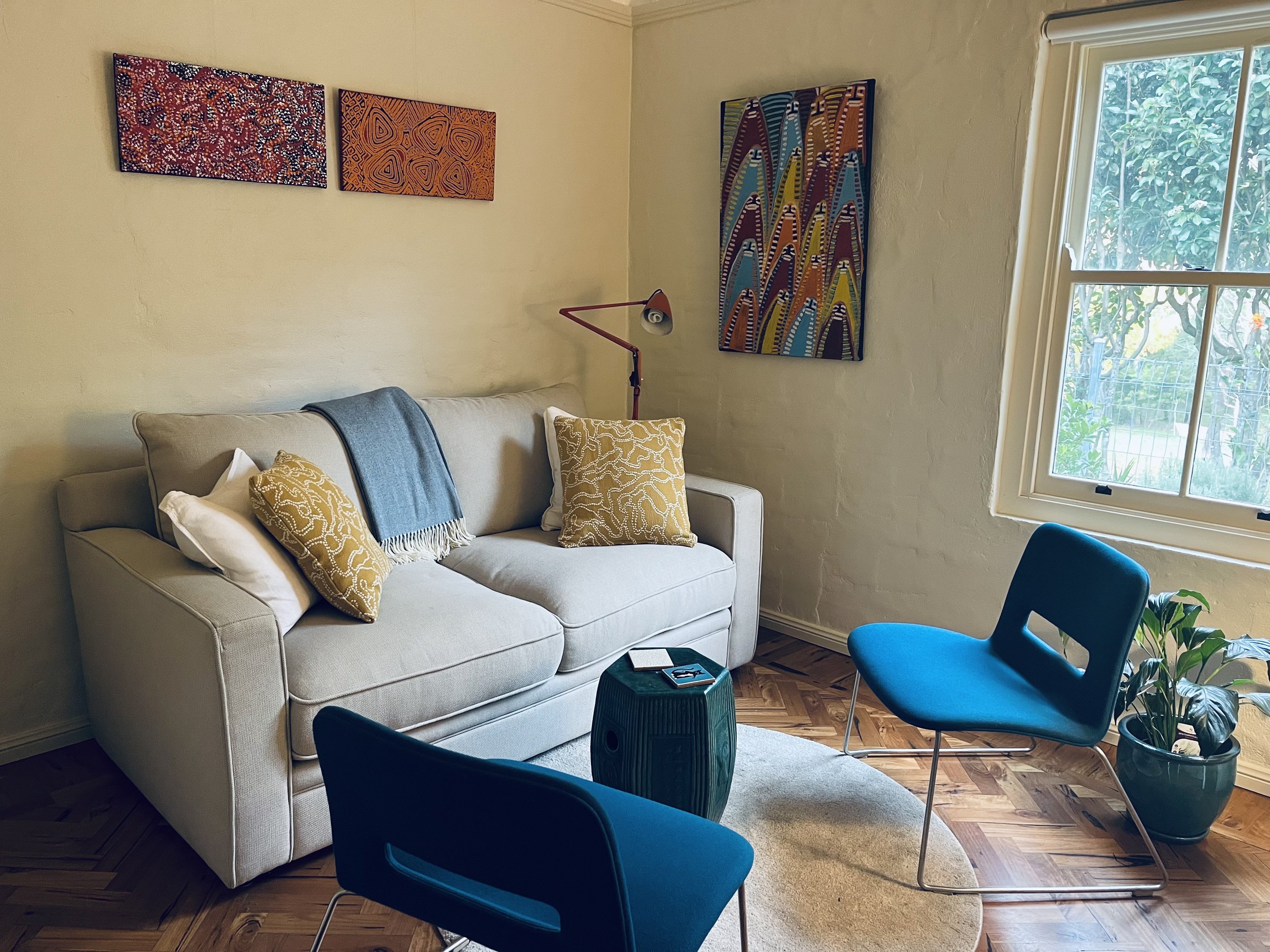 Living room with beige sofa decorated with yellow patterned pillows and a throw blanket, a brown wooden floor, a blue chair, a small table, a potted plant, artwork on the walls, a window with greenery outside.