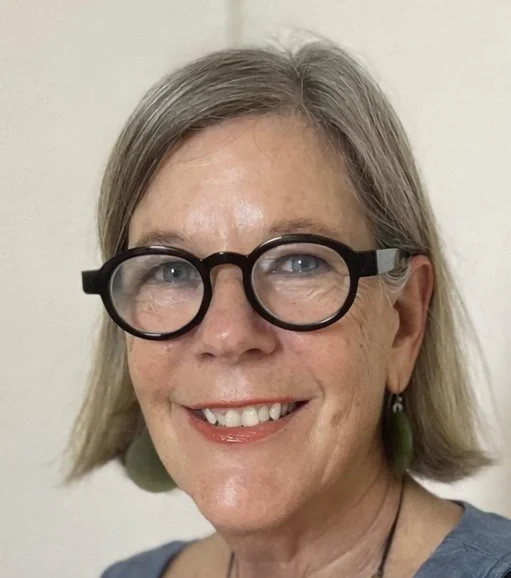 A smiling woman with gray hair, large black glasses, and green earrings, wearing a blue top.