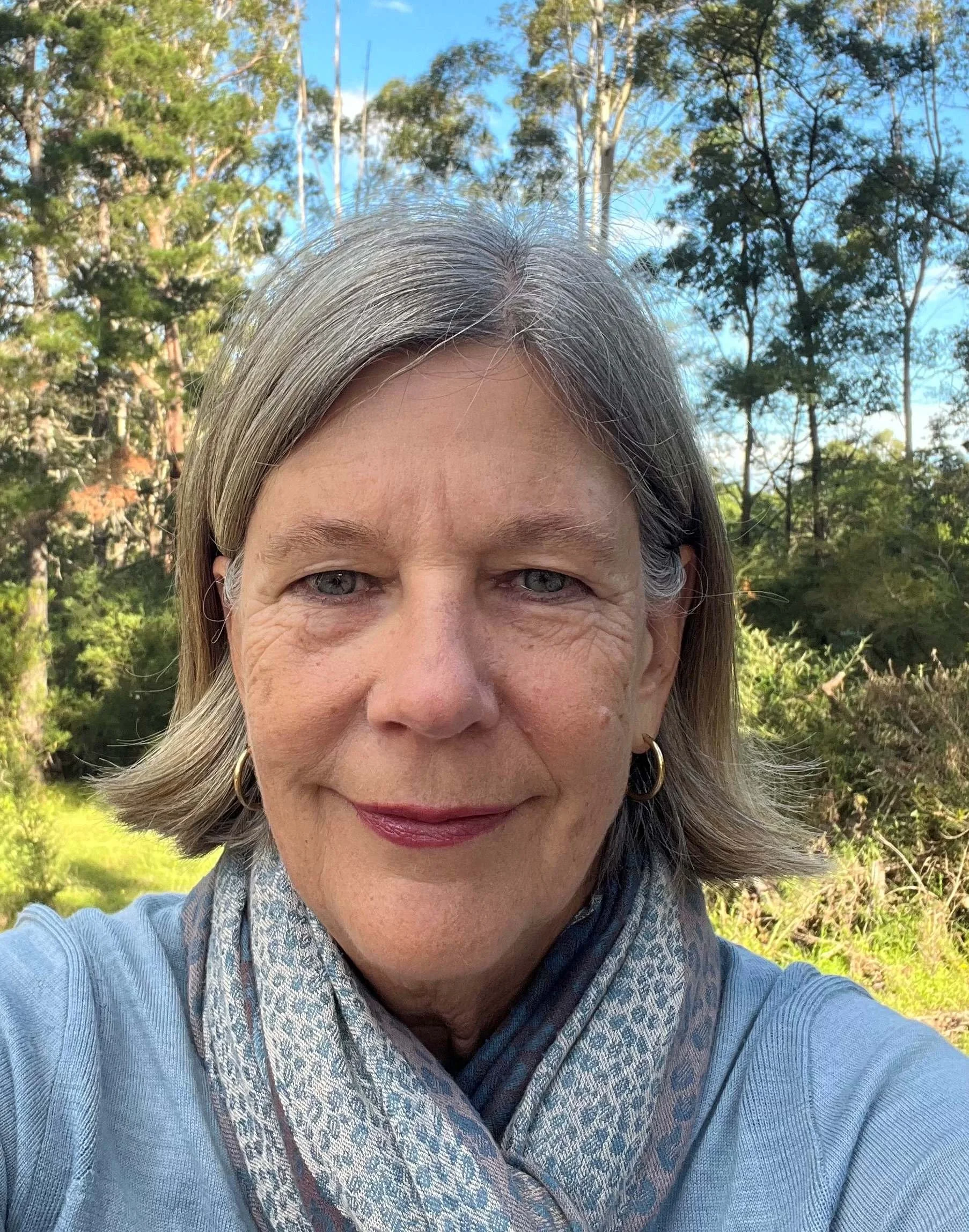 Close-up of a smiling woman with gray hair, wearing earrings and a scarf, outdoors with trees and a blue sky in the background.