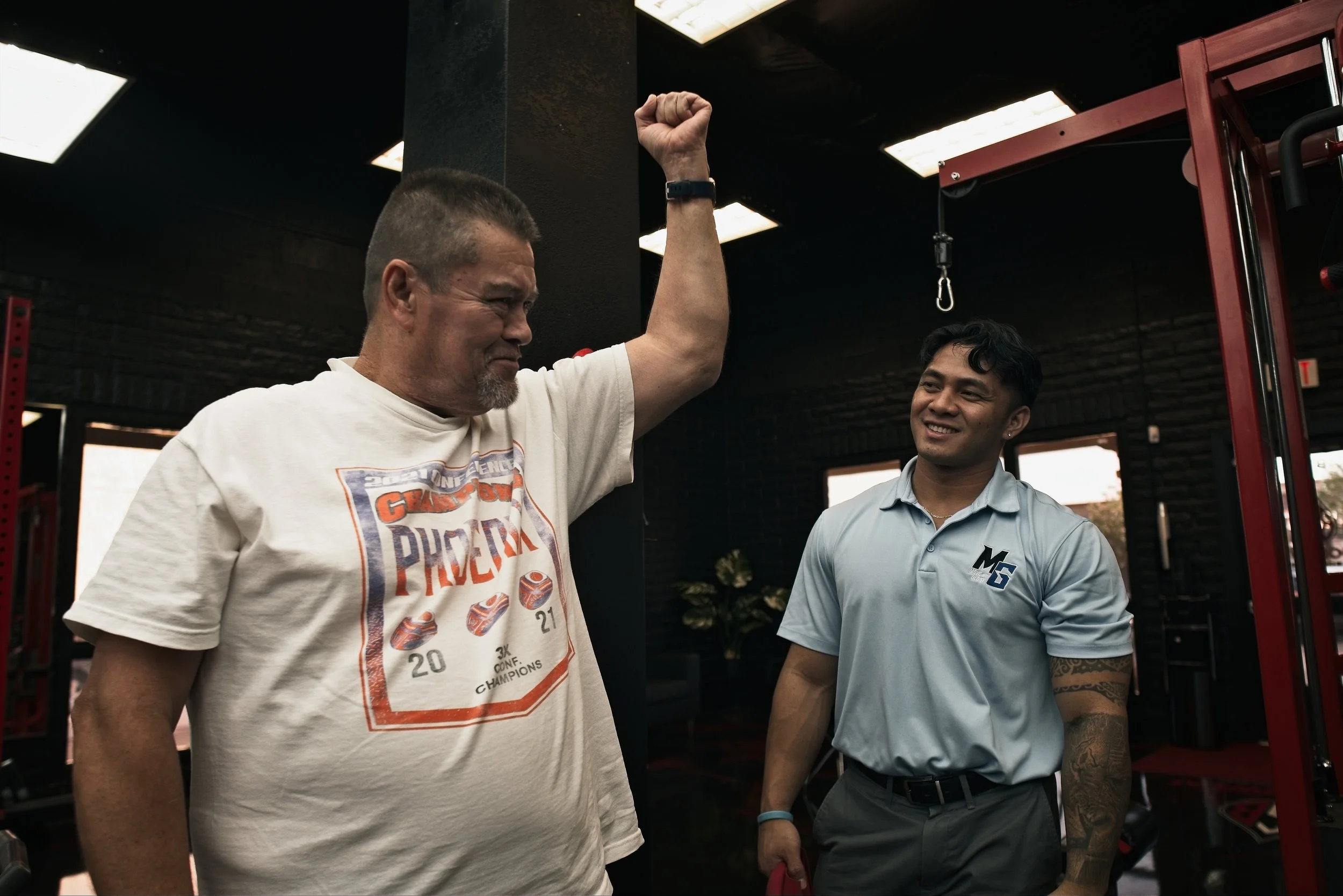 Two men in a gym, one with a muscular build and short hair raising his fist, smiling, the other standing beside him, wearing a light blue polo shirt and smiling.