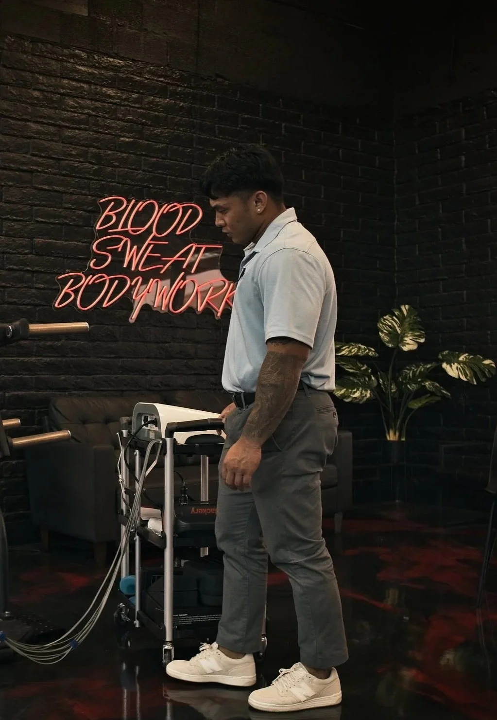 A young man with tattoos and a white polo shirt standing next to medical equipment in a gym or wellness center. A neon sign reading 'Blood Sweat Bodywork' is illuminated on a black brick wall behind him, with a couch and potted plant also visible.