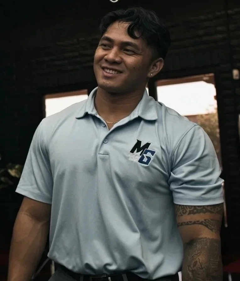 Young man with dark curly hair smiling, wearing a light gray polo shirt with Columbia logo, standing indoors with a dark background and windows.