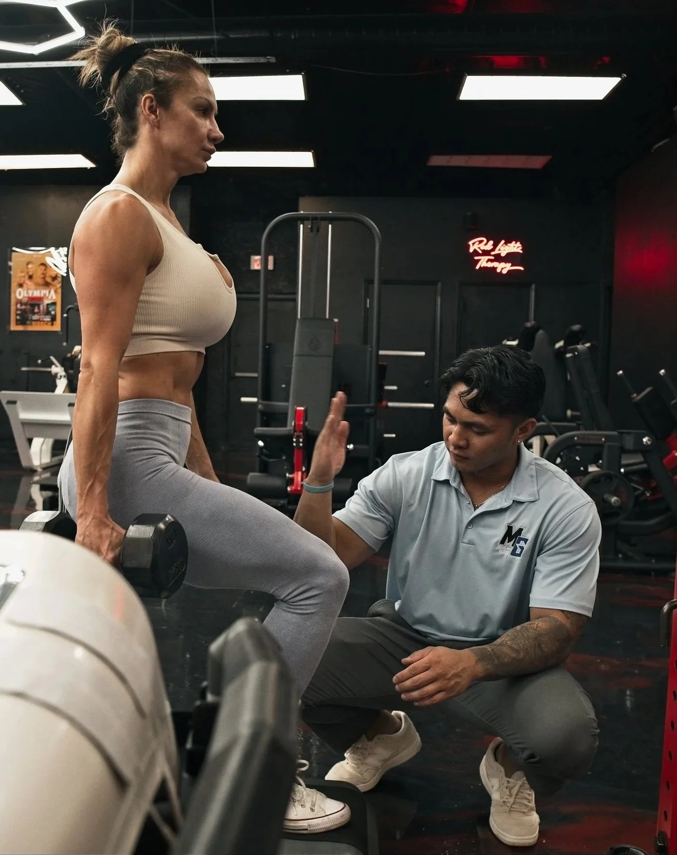 A woman performing a squat with dumbbells while a trainer assists her at a gym.