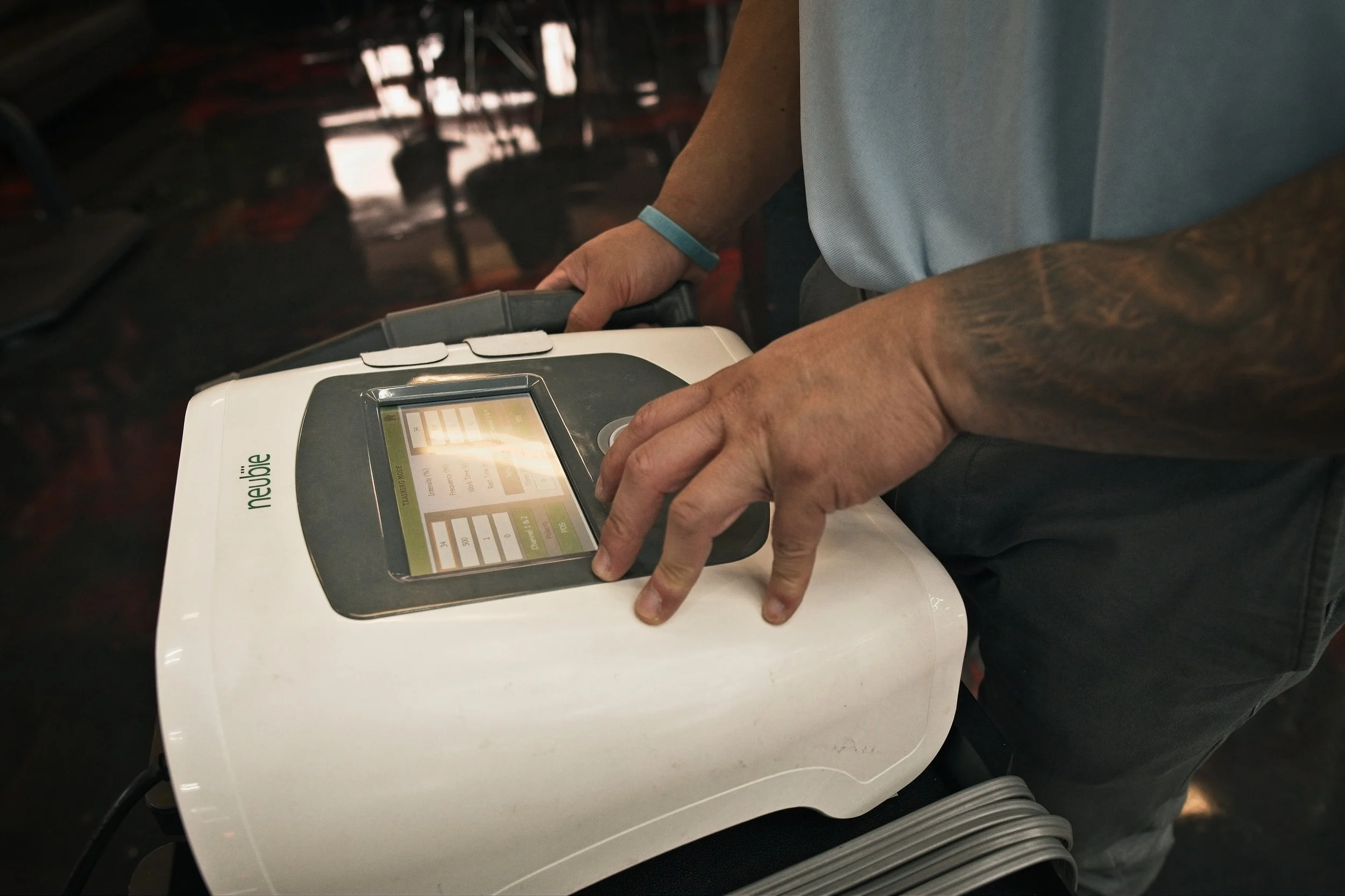 A person, with a tattooed arm, using a medical or fitness device with a touchscreen interface in a gym or clinical setting.