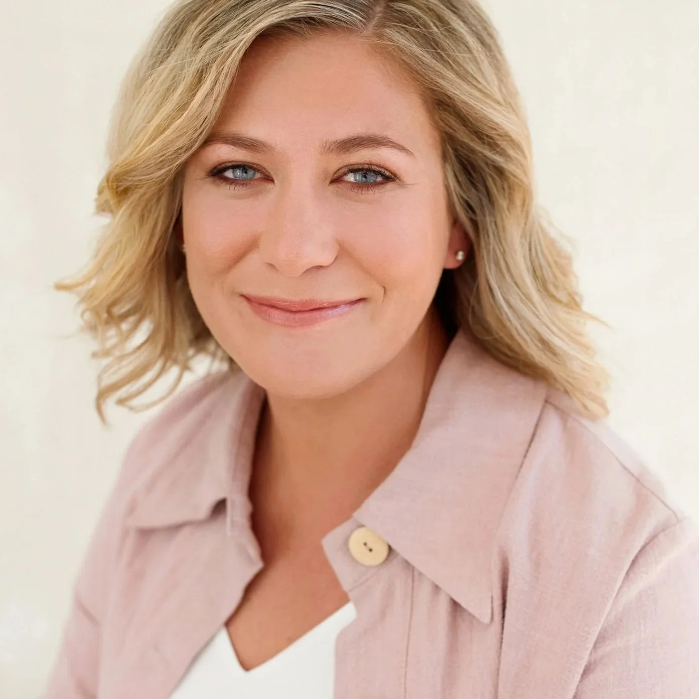 Close-up portrait of a woman with shoulder-length blonde hair, blue eyes, wearing a light pink button-up shirt, smiling softly against a neutral background.