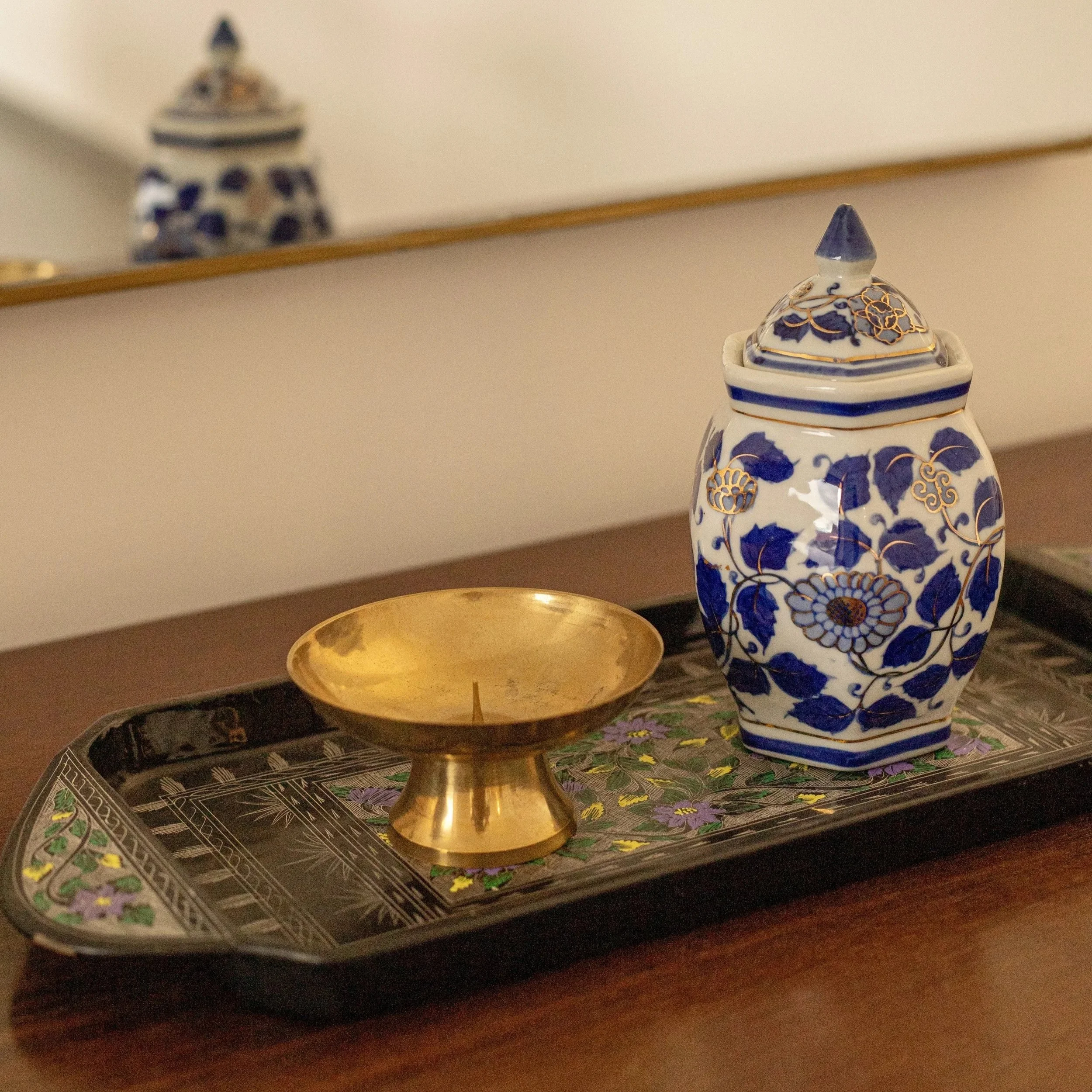 Une jarre en porcelaine blanche avec un motif de fleurs et de feuilles bleues et or, une coupelle en or, et un plateau décoré de motifs floraux, sur une table en bois.