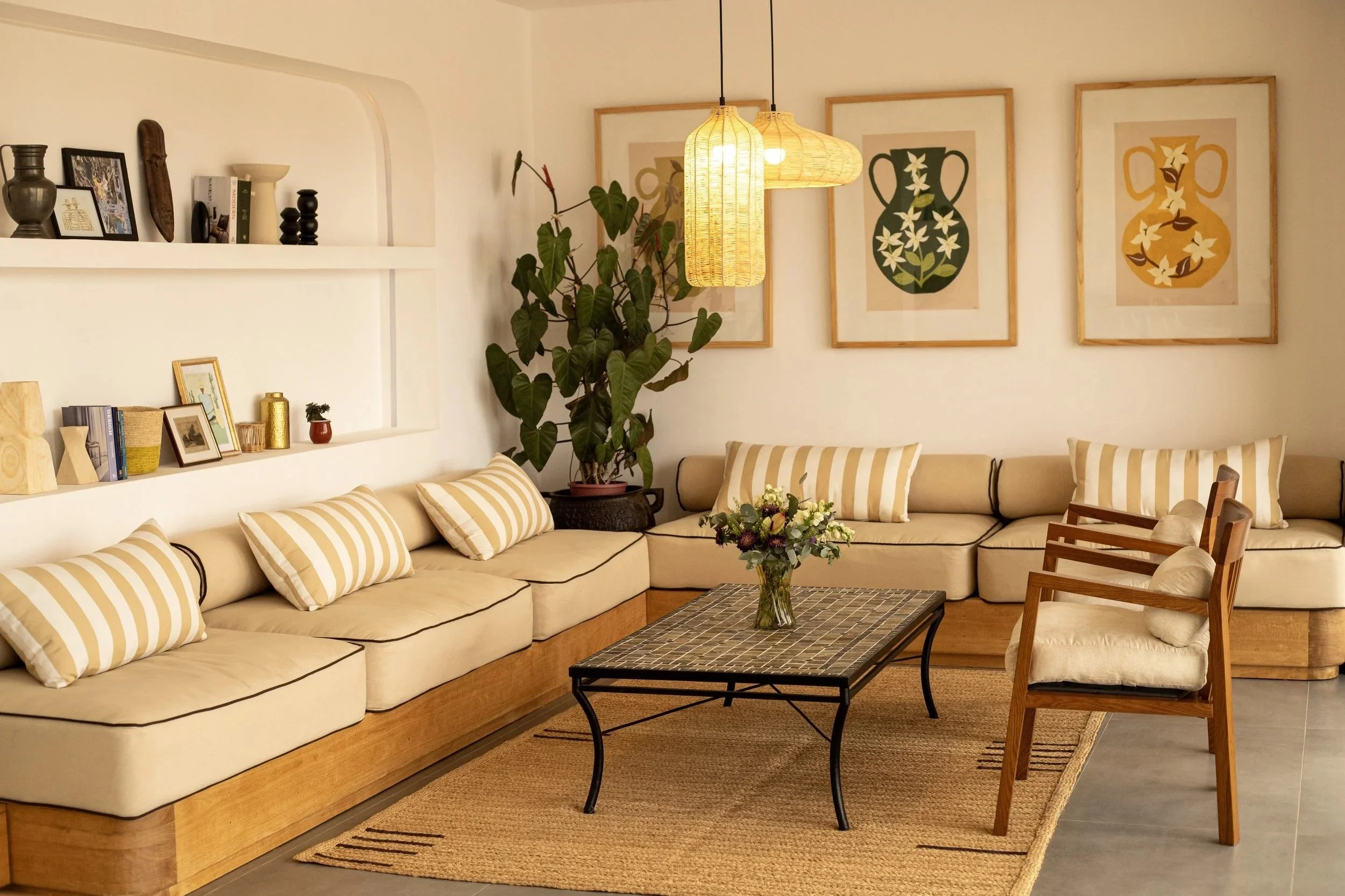 Salon avec canapé beige, coussins à rayures jaunes et blanches, tableau de vases avec fleurs, chandelier en papier suspendu, table basse avec bouquet de fleurs, fauteuils en bois et tissu beige, tapis en raphia.