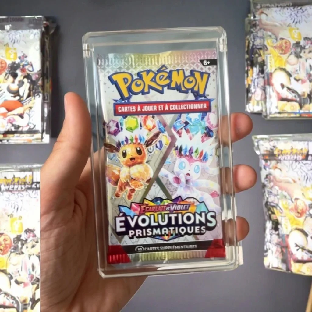 A Pokémon card pack of Eeveelutions Prismatic with illustrations of Eevee and its evolutions, held in a person's hand with other Pokémon card packs on display in the background.