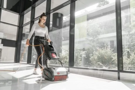 Woman using a floor cleaning machine in a modern, glass-walled building.