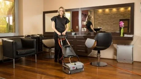 A woman cleaning a wooden floor with a floor buffer in a modern living room with black chairs, a mirror, and a brick wall in the background.