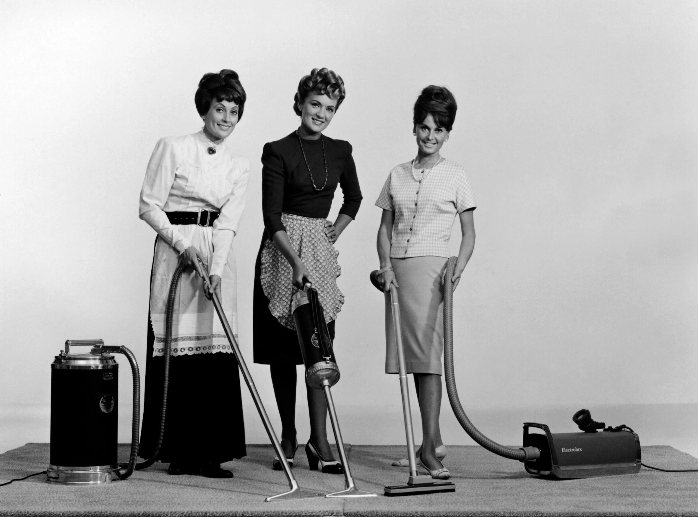Three women in 1960s attire holding vacuum cleaners, standing on a carpeted stage with a plain background.