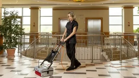 A woman is operating a floor cleaning machine in a spacious, well-lit indoor area that appears to be a hotel or office lobby.