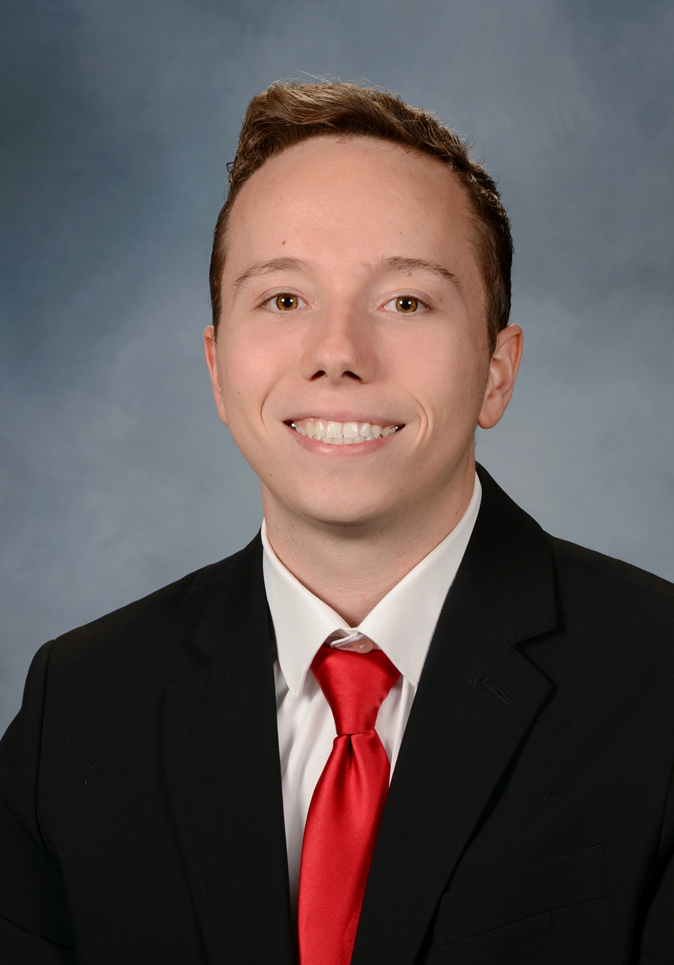 A young man in a black suit, white shirt, and red tie, smiling against a gray background.